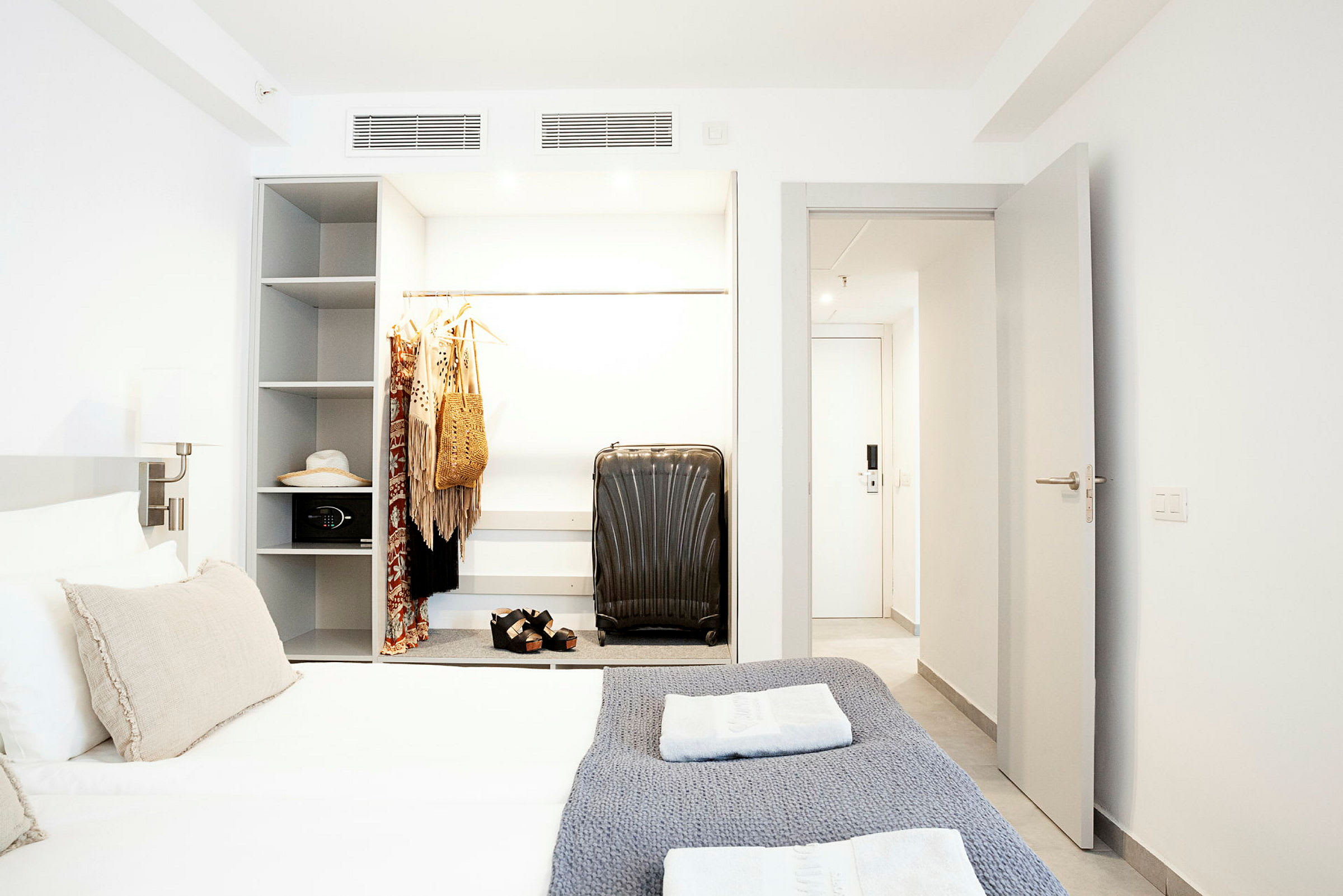Hotellihuone, jossa sänky, vaatekaappi roikkuvilla vaatteilla ja matkalaukku. (Tekoälyn luoma teksti)