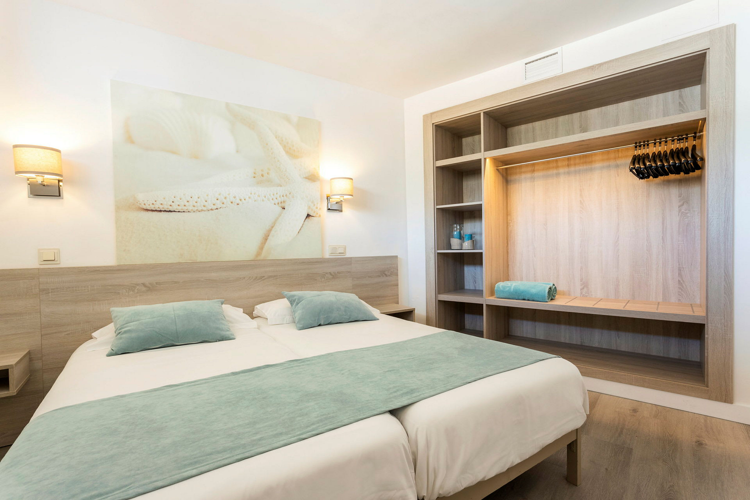 Hotellihuone, jossa on parivuode, siniset vuodevaatteet, seinälamput ja avoin vaatekaappi. (Tekoälyn luoma teksti)