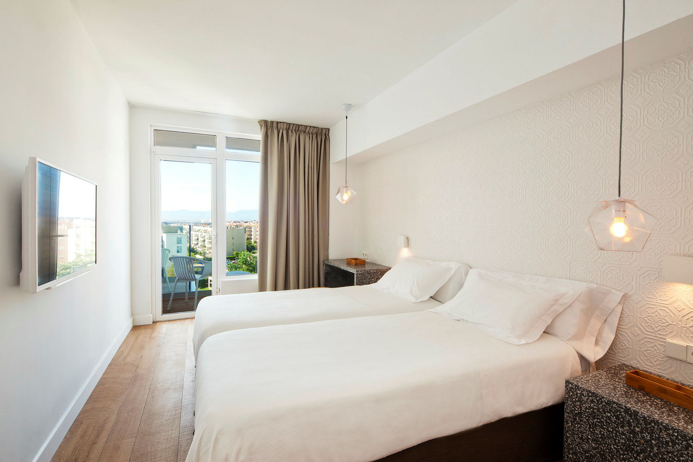Hotellihuone, jossa on kaksi erillistä sänkyä, valkoiset vuodevaatteet, seinälle asennettu televisio ja näkymä kaupunkiin lasioven kautta. (Tekoälyn luoma teksti)