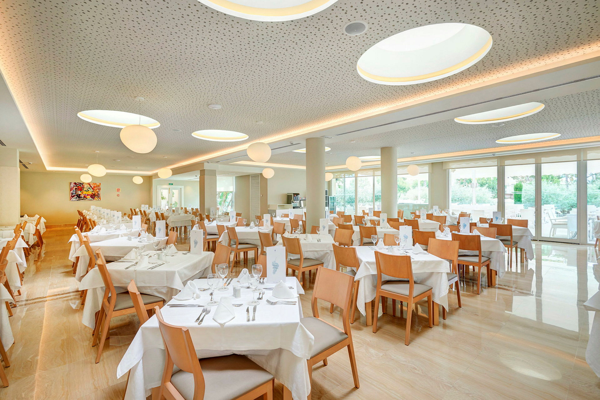 Ravintola, jossa on useita pöytiä ja puisia tuoleja, suuret ikkunat ja kattovalaisimet. (Tekoälyn luoma teksti)