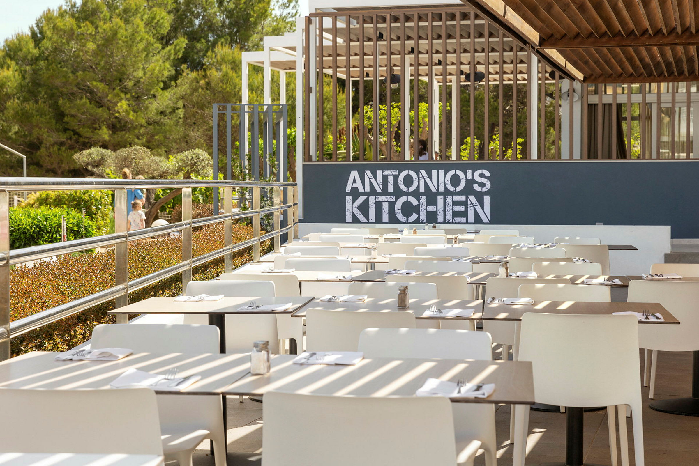 Ulkoterassi, jossa on valkoisia tuoleja ja katettuja pöytiä kyltin edessä, joka lukee 'ANTONIO'S KITCHEN'. (Tekoälyn luoma teksti)