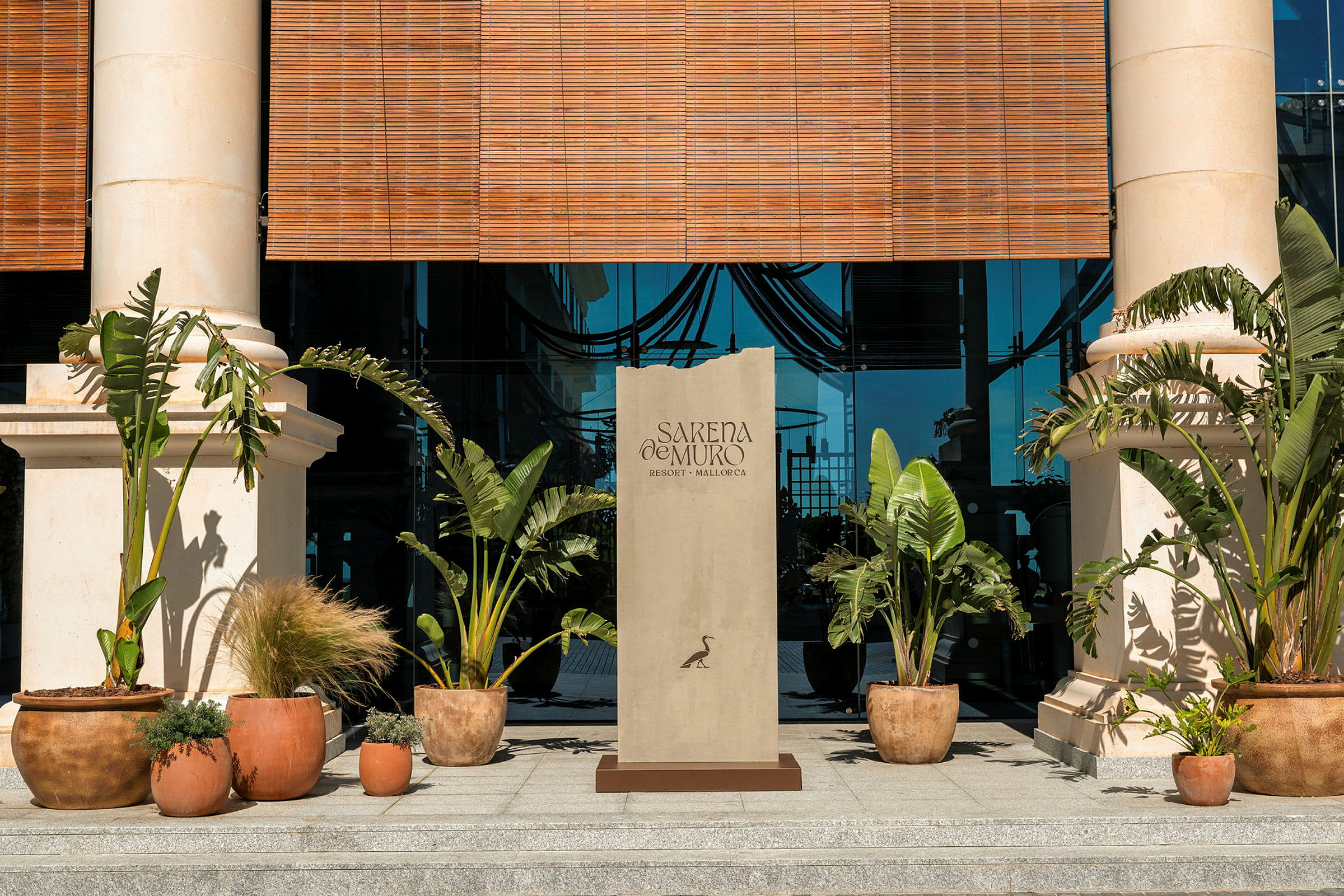 Sisäänkäynti rakennukseen, jossa on kaksi pylvästä, vihreitä kasveja ruukuissa ja kyltti, jossa lukee "Sarena de Muro Resort Mallorca". (Tekoälyn luoma teksti)