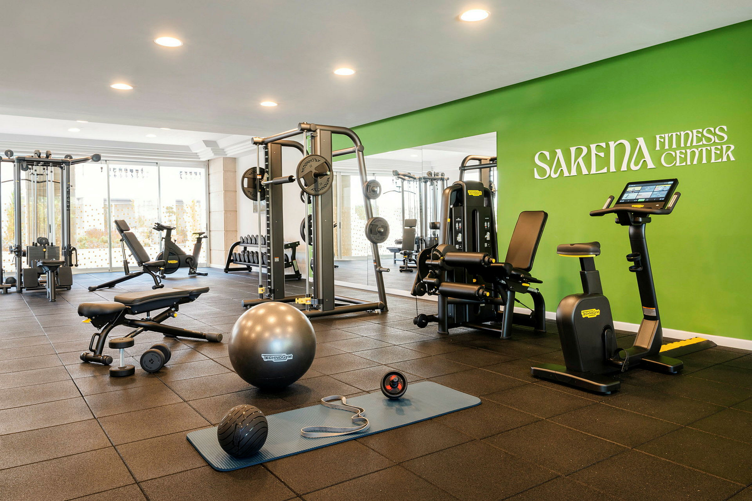 Kuntosali, jossa on erilaisia kuntoilulaitteita ja välineitä, kuten kuntopyörä, käsipainot ja harjoittelumatto, sekä vihreä seinä, jossa lukee Sarena Fitness Center. (Tekoälyn luoma teksti)