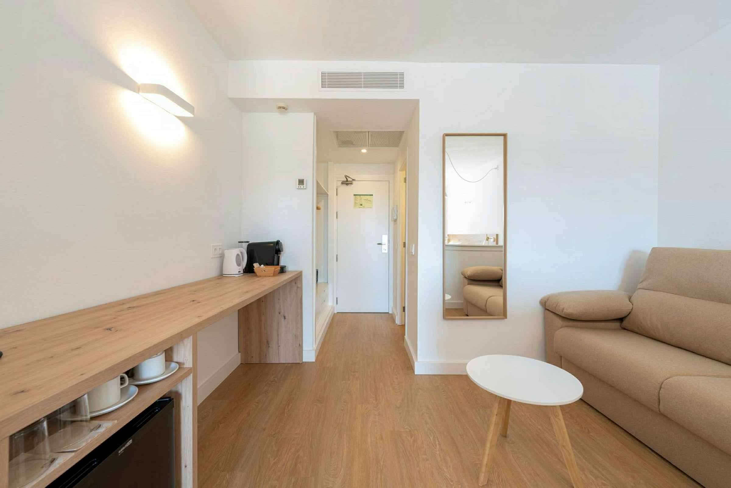 Moderni hotellihuone, jossa on sohva, peili, pieni pöytä ja työtila vedenkeittimellä ja kahvinkeittimellä. (Tekoälyn luoma teksti)