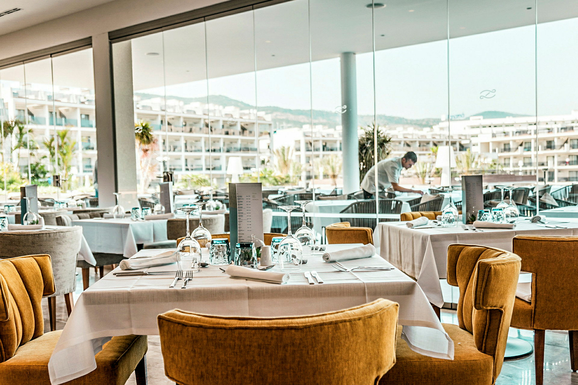 Moderni ravintola, jossa on katettuja pöytiä ja tuoleja sekä ikkunoita, joista on näkymä hotellialueelle. (Tekoälyn luoma teksti)