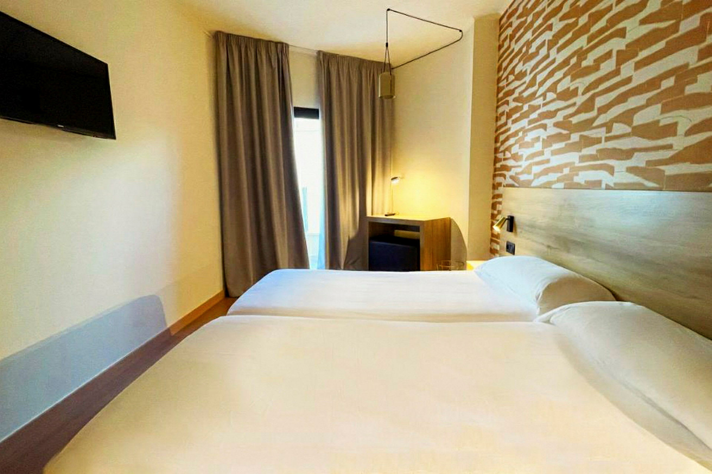 Kaksi yhden hengen sänkyä hotellihuoneessa, jossa on kuvioitu pääty ja televisio seinällä. (Tekoälyn luoma teksti)