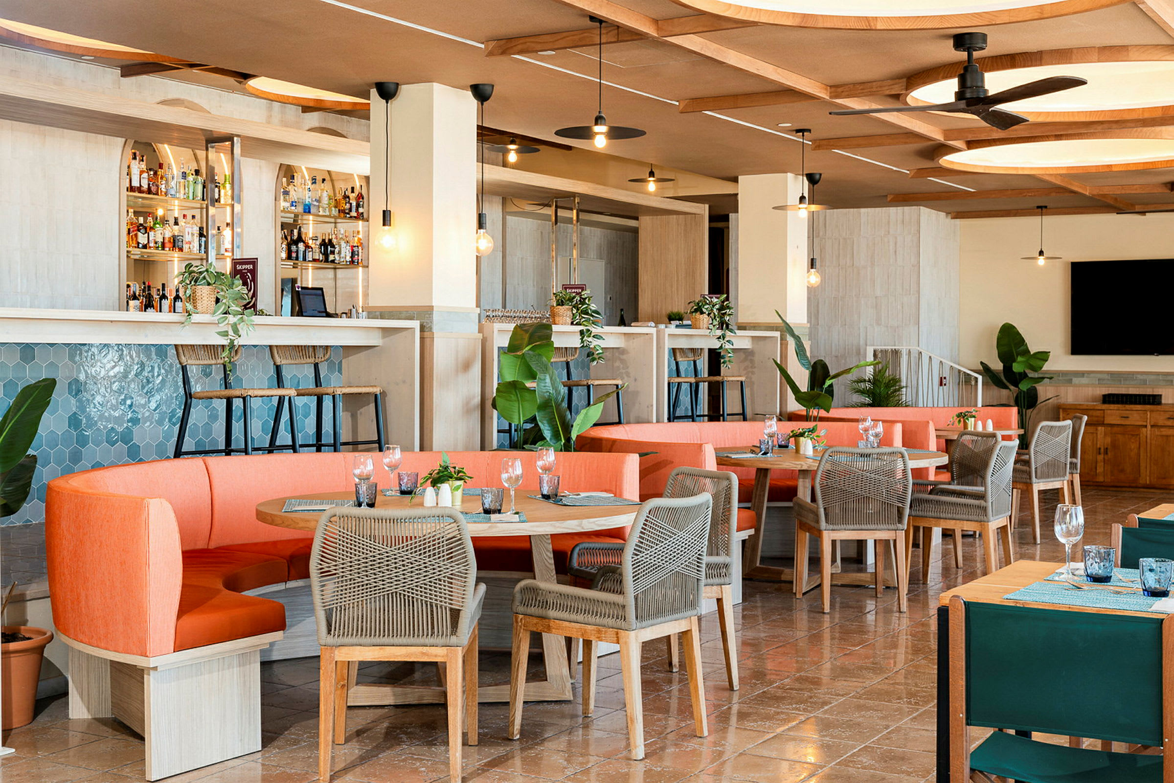 Moderni ravintola, jossa on kutsuva sisustus, oransseja sohvia ja harmailla tuoleilla varustettuja pöytiä sekä baaritiski ja vihreitä kasveja. (Tekoälyn luoma teksti)