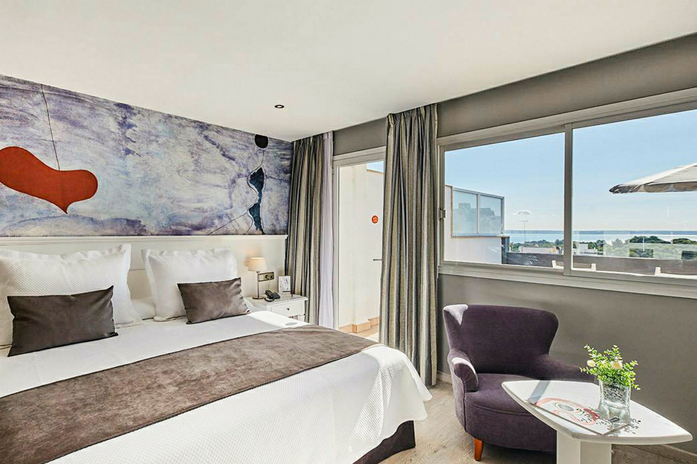 Moderni hotellihuone, jossa on parivuode, värikäs seinämaalaus ja suuret ikkunat merinäköalalla. (Tekoälyn luoma teksti)