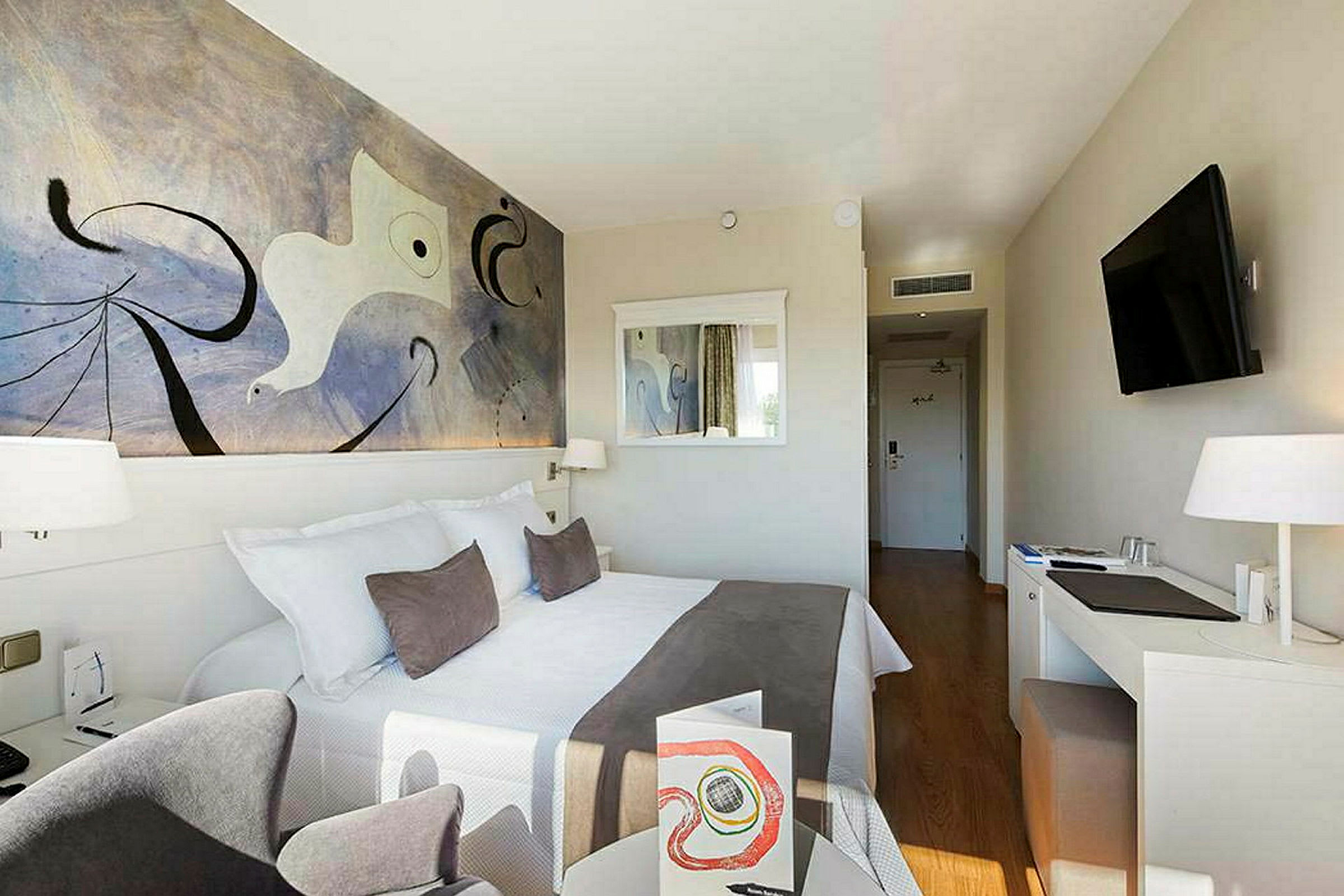 Hotellihuone, jossa parisänky, seinätaidetta, peili, kirjoituspöytä ja televisio. (Tekoälyn luoma teksti)