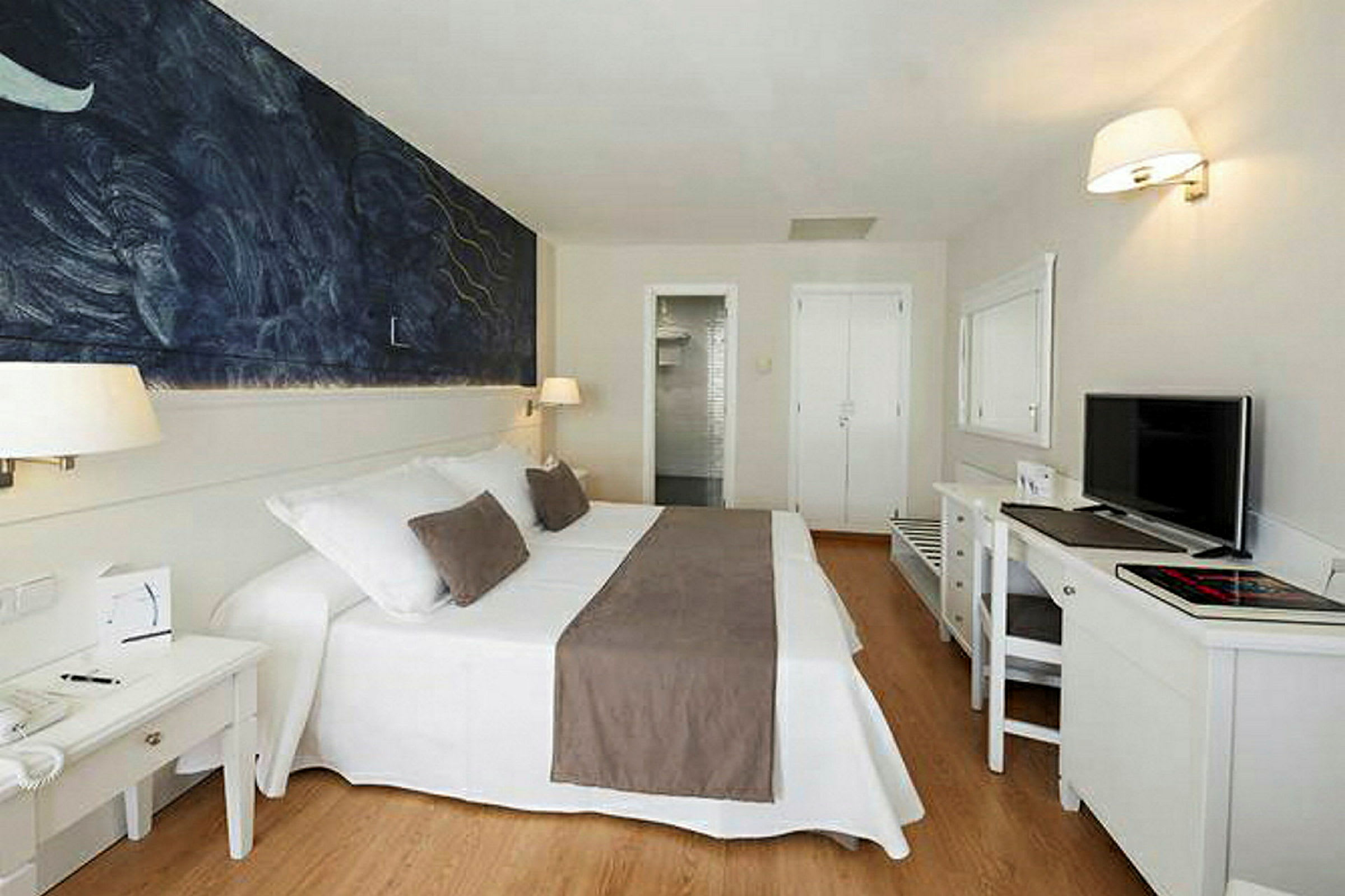 Hotellihuone, jossa on parivuode valkoisin ja ruskein vuodevaattein, seinällä maalaus ja työpöytä, jolla on televisio. (Tekoälyn luoma teksti)