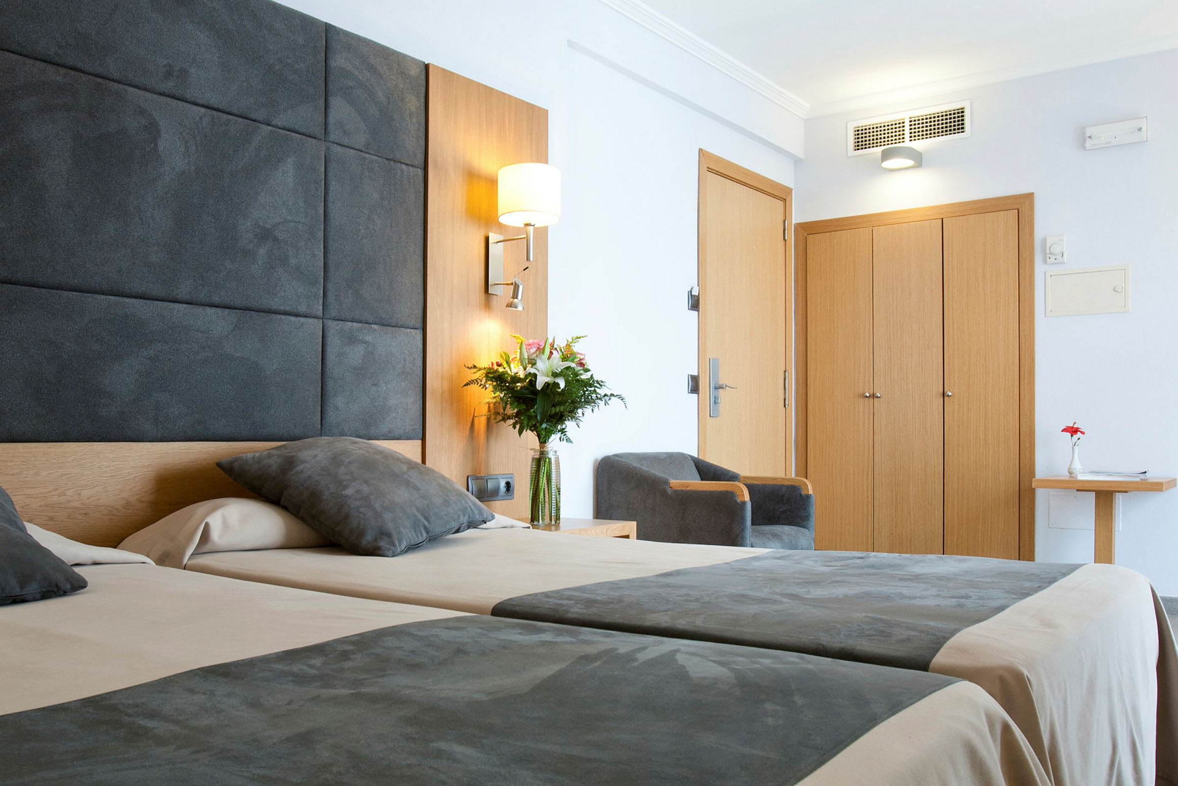 Kuva modernista hotellihuoneesta, jossa on kaksi vuodetta, harmaat päiväpeitot, tuoli, puuvaatekaappi ja pöydällä oleva kukkavaasi. (Tekoälyn luoma teksti)