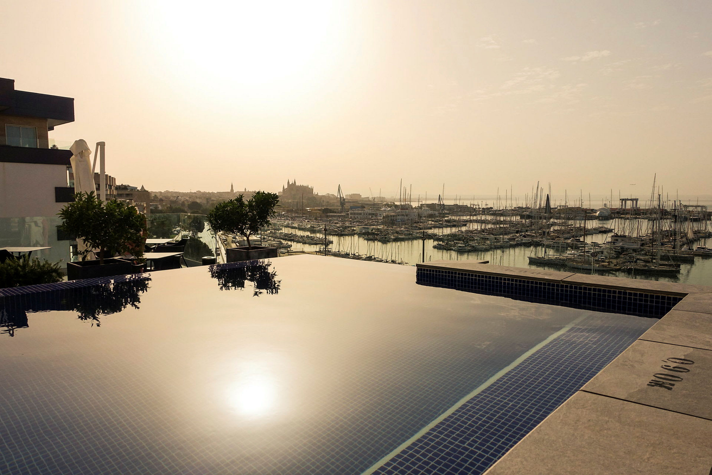 Kuva näkymästä venesatamaan, jossa on useita purjeveneitä, nähtynä uima-altaalta, joka heijastaa auringonlaskun. (Tekoälyn luoma teksti)