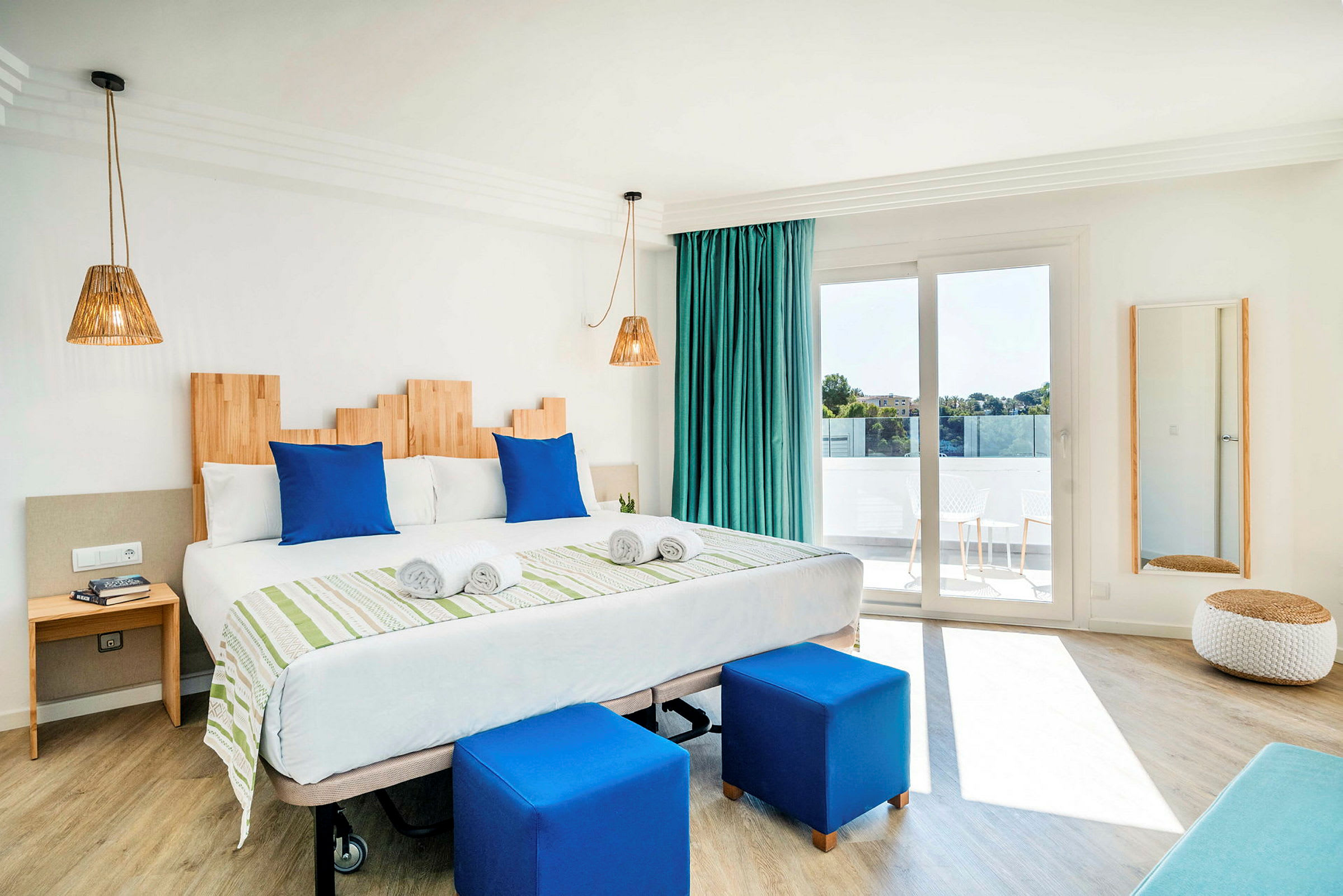 Hotellihuone, jossa sänky, siniset tyynyt ja jakkarat, vihreät verhot ja parveke. (Tekoälyn luoma teksti)