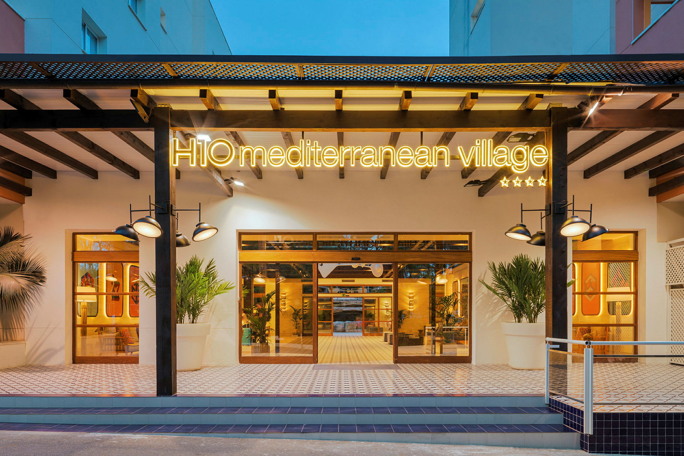 H10 Mediterranean Village -hotellin sisäänkäynti, jossa on valaistu kyltti, suuret ikkunat ja moderni muotoilu. (Tekoälyn luoma teksti)