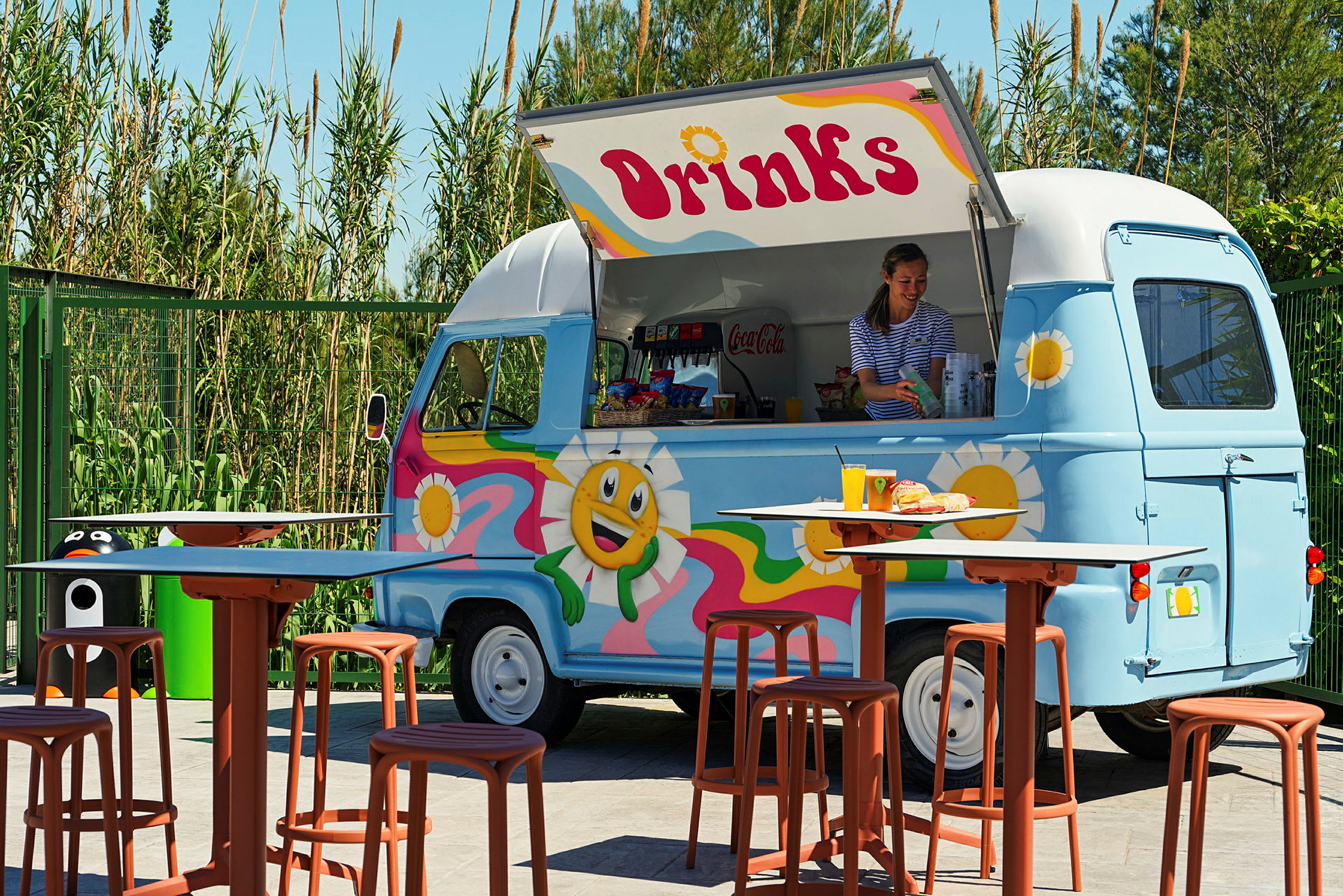 Värikäs ruokarekka, jossa lukee 'Drinks', tarjoilee juomia ja välipaloja. Rekan edessä on pöytiä ja tuoleja. (Tekoälyn luoma teksti)