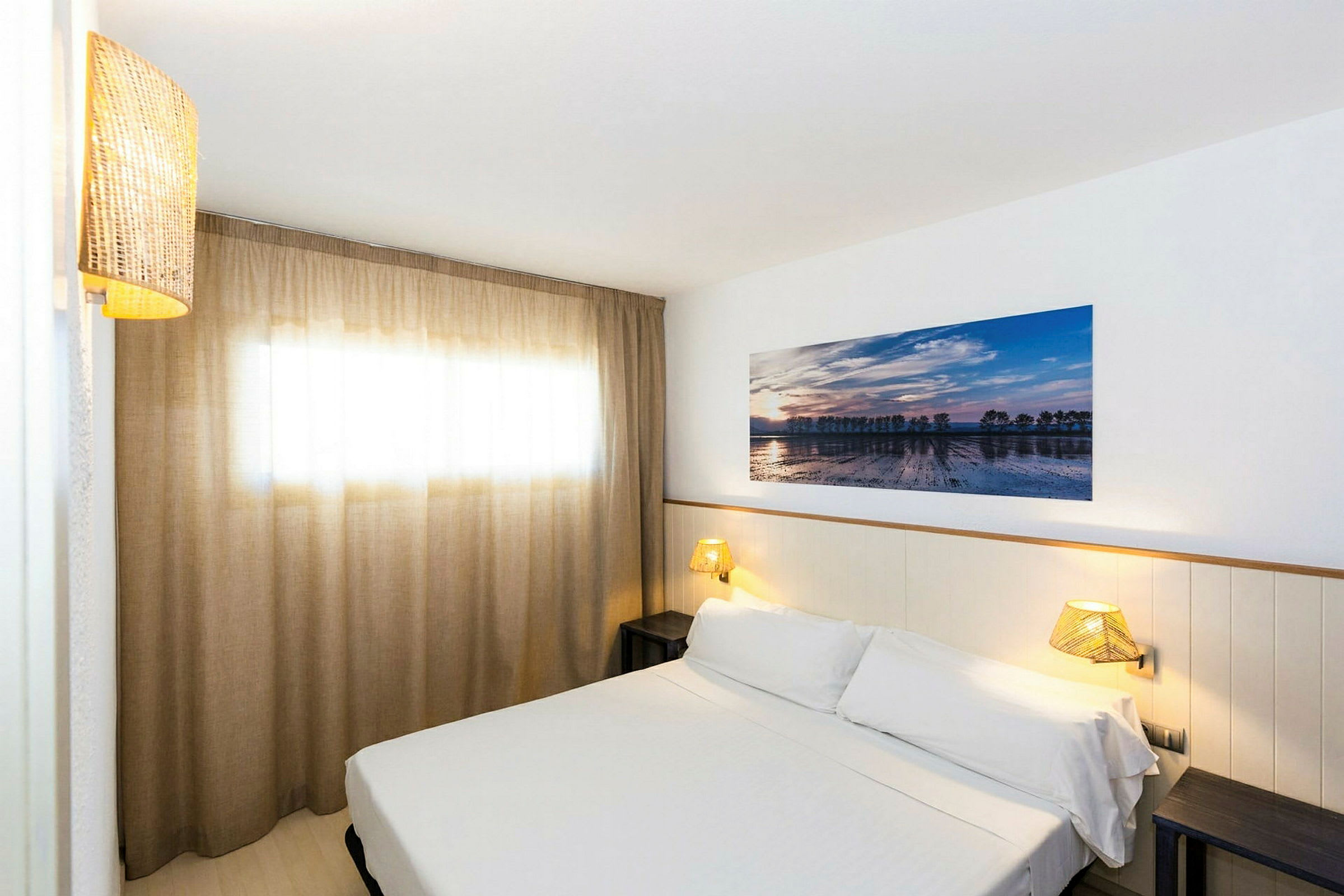 Hotellihuone, jossa on valkoisilla vuodevaatteilla vuorattu sänky, seinävalaisin ja seinälle ripustettu taideteos. Ikkuna, jossa on tyylikkäät beiget verhot. (Tekoälyn luoma teksti)