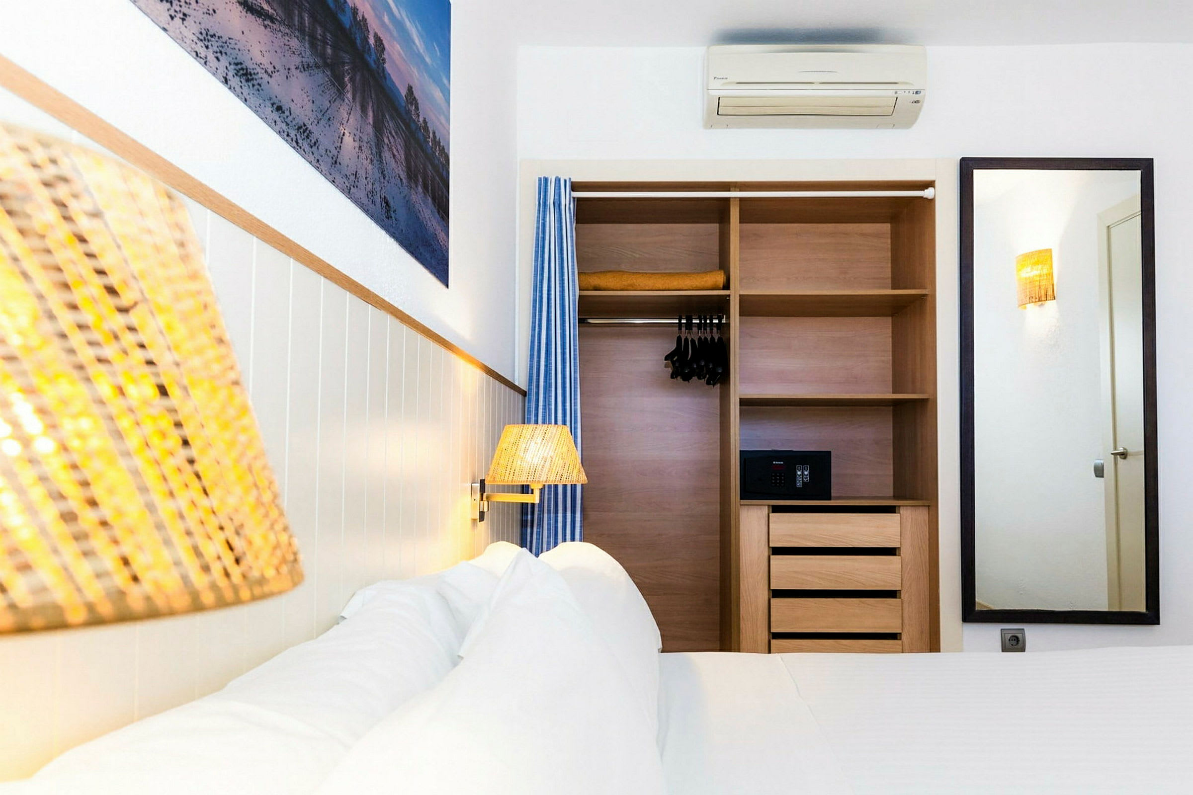 Hotellihuone, jossa on sänky ja kiinteä vaatekaappi. Kaappi on auki ja siinä on hyllyjä, henkareita ja tallelokero. Seinällä on peili ja taulu. (Tekoälyn luoma teksti)