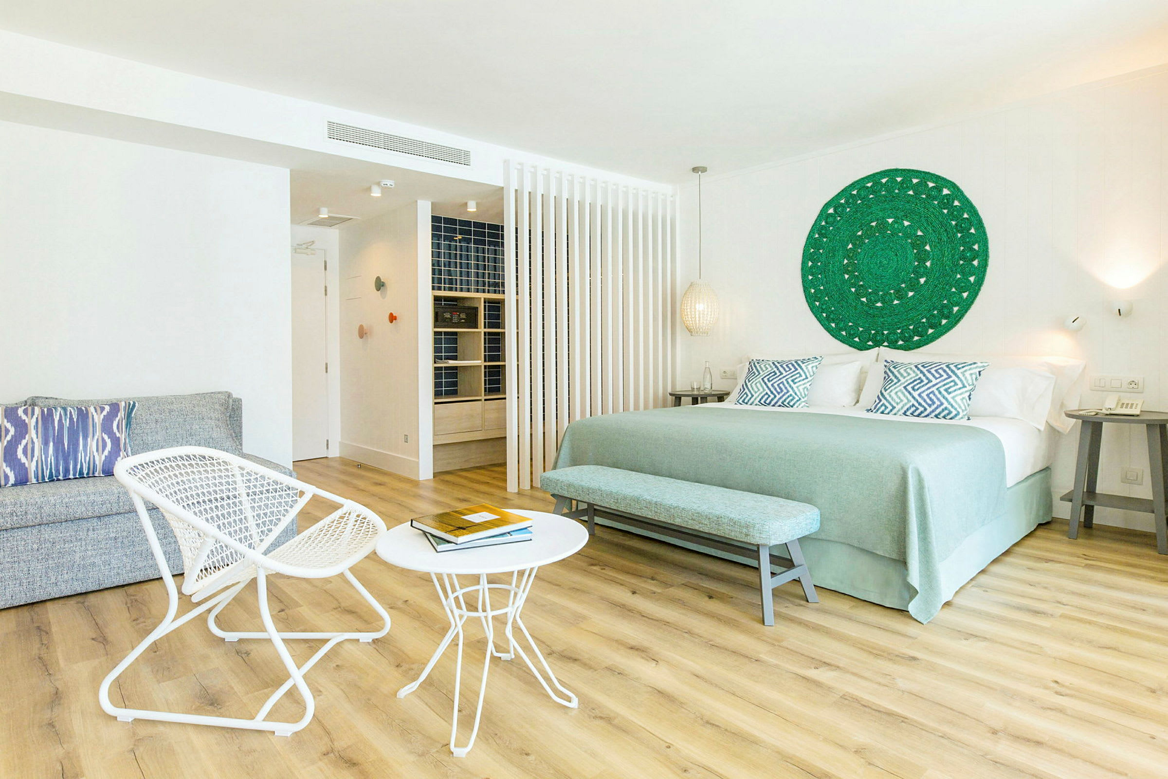 Valoisa hotellihuone, jossa on iso sänky, kaksi pientä pöytää ja kaksi tuolia sekä koristeellinen seinäkoriste. (Tekoälyn luoma teksti)