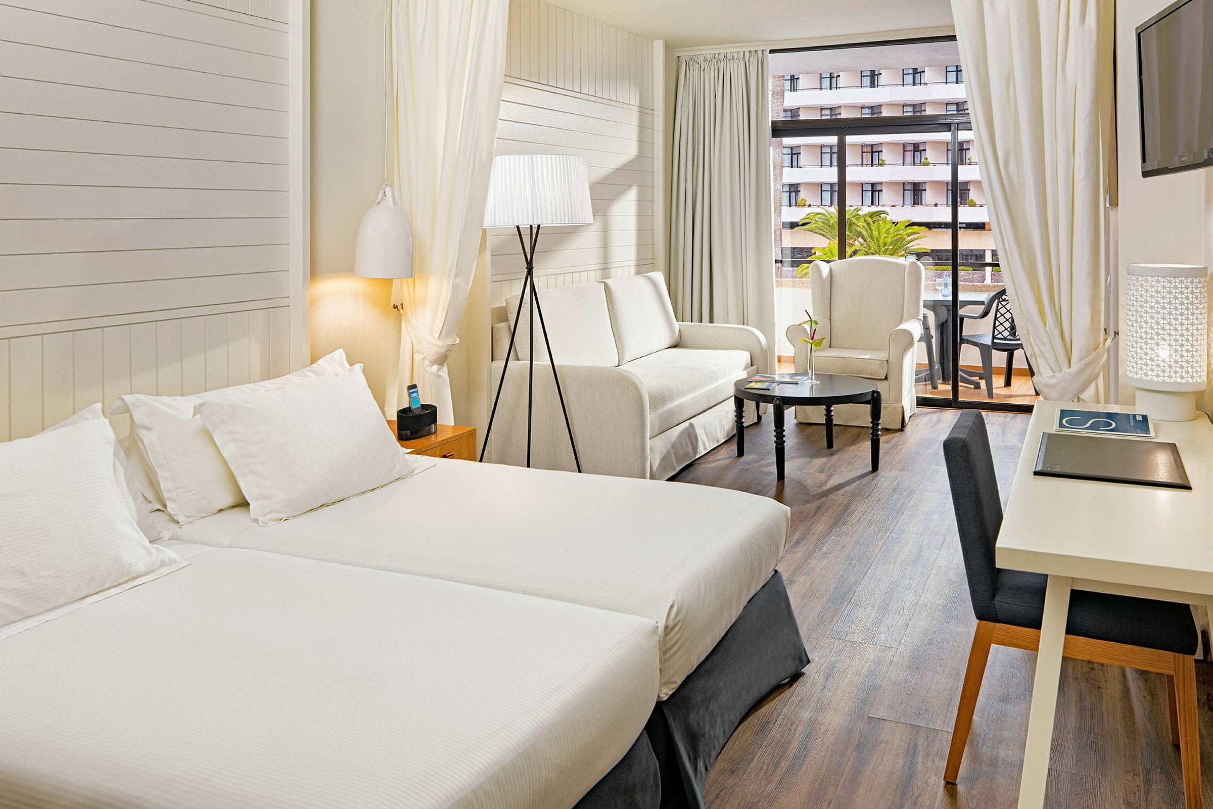 Hotellihuone, jossa on kaksi erillistä vuodetta, istuinryhmä sohvan ja nojatuolin kanssa sekä pöytä. Lisäksi huoneessa on kirjoituspöytä ja parveke. (Tekoälyn luoma teksti)