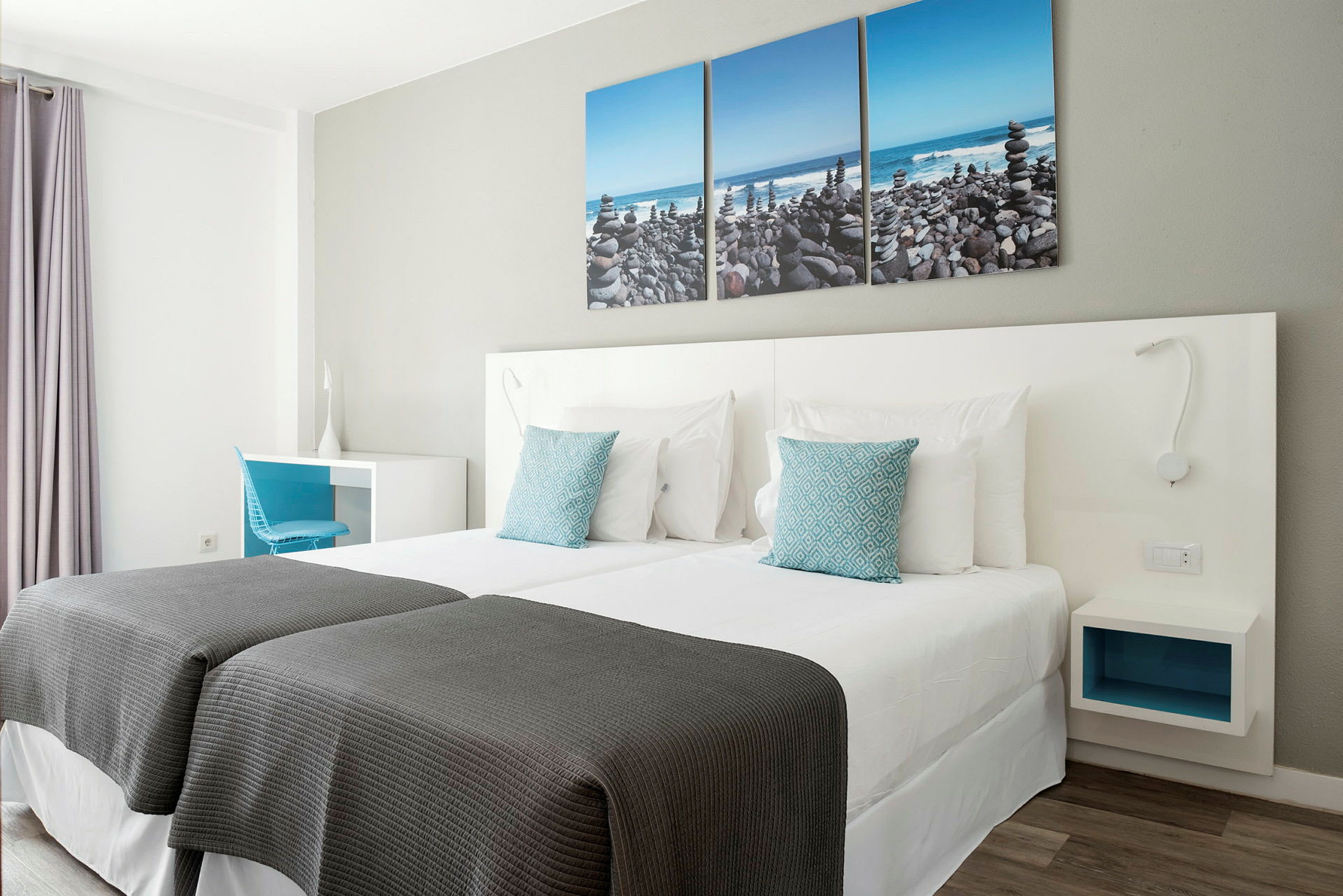 Hotellihuone, jossa on kaksi yksittäissänkyä, valkoiset vuodevaatteet ja siniset koristeelliset tyynyt. Seinällä on kolmen osan taulu, joka esittää kivirantaa. (Tekoälyn luoma teksti)