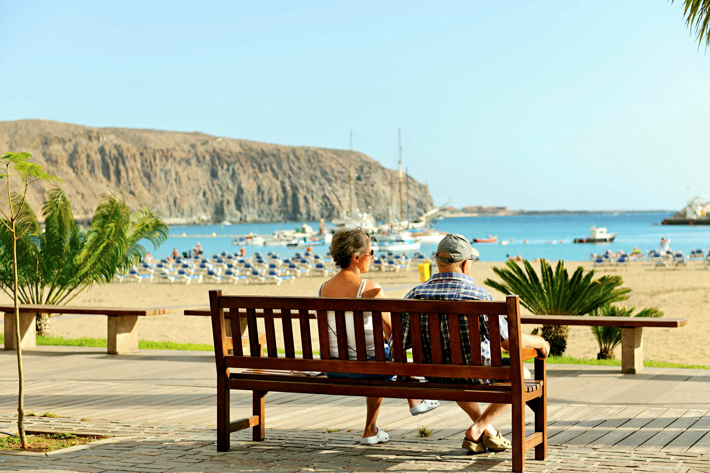 Kaksi henkilöä istuu penkillä katsellen hiekkarantaa ja merta, taustalla veneitä ja kallioita. (Tekoälyn luoma teksti)