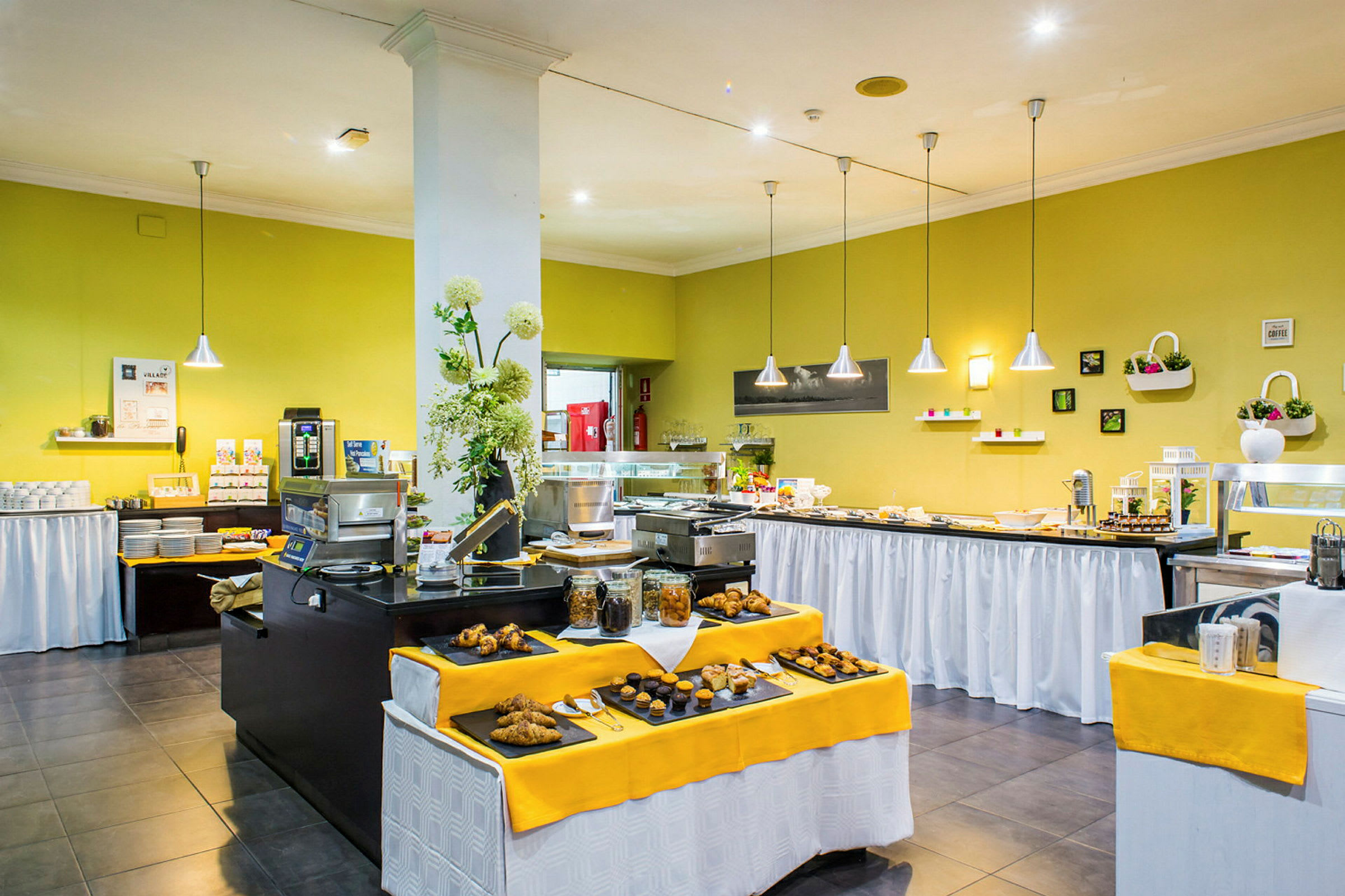 Buffet-pöydät, joissa on erilaisia ruokia, koristeltu keltaisilla pöytäliinoilla ja ympäröity vihreillä seinillä ja riippuvilla valoilla. (Tekoälyn luoma teksti)