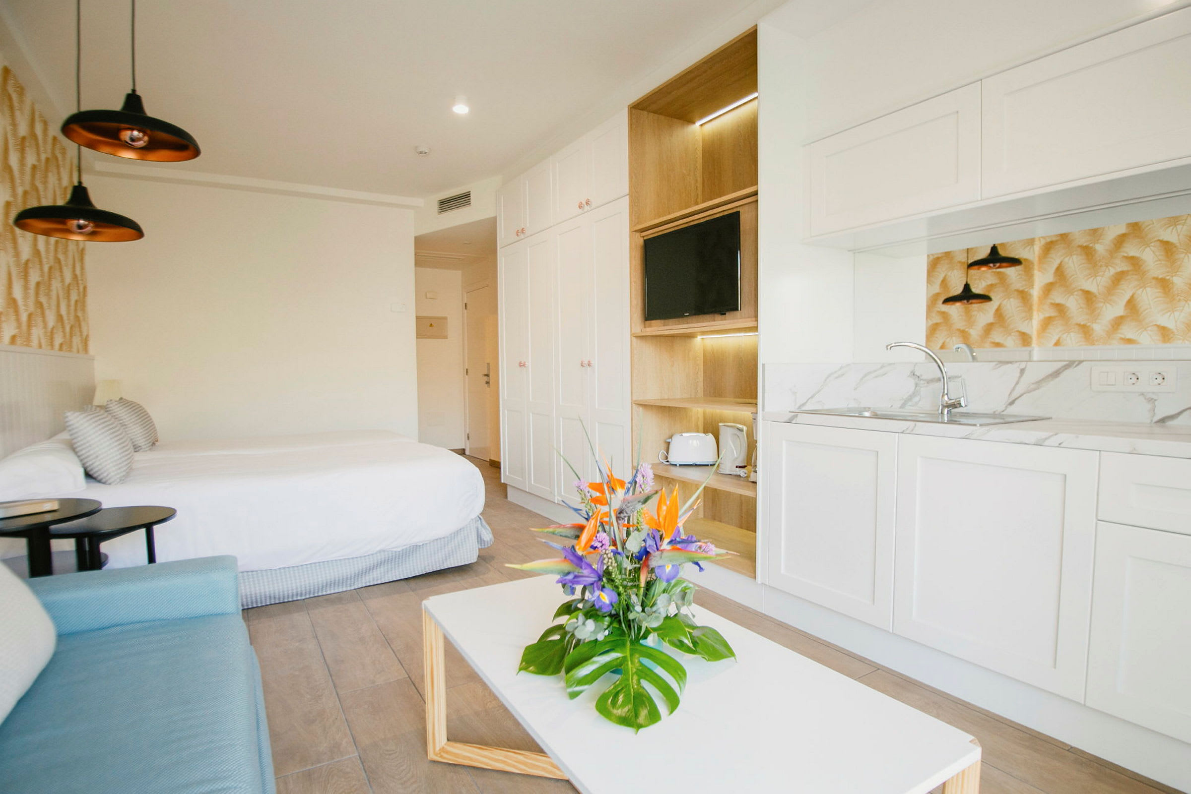 Moderni hotellihuone, jossa on parisänky, sohva, pieni keittiö ja pöytä kukkakimpulla. (Tekoälyn luoma teksti)