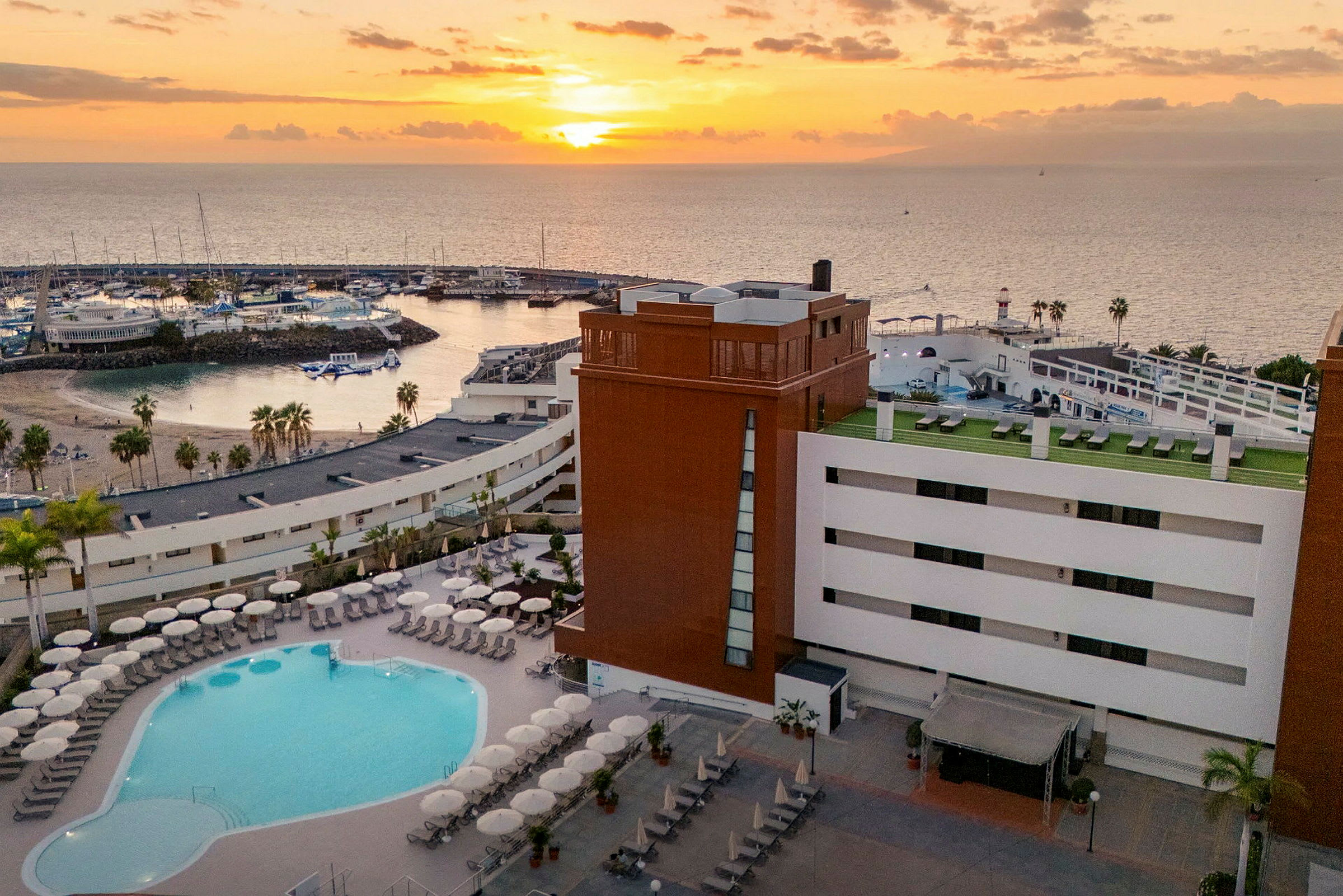 Kuva hotellista, jonka etualalla on uima-allasalue ja taustalla satama auringonlaskun aikaan. (Tekoälyn luoma teksti)