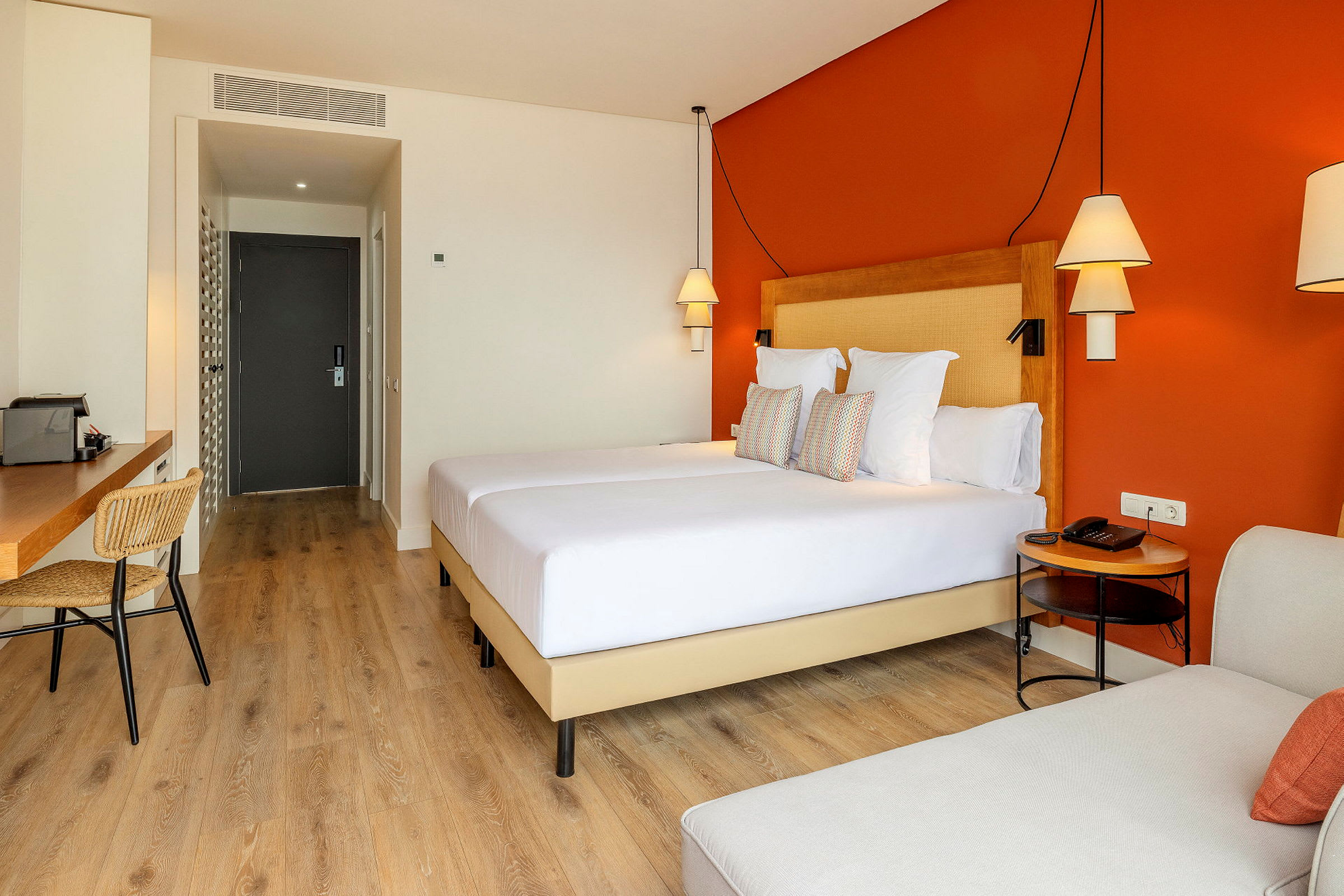 Hotellihuone, jossa oranssi seinä, parisänky, työpöytä ja tuoleja. (Tekoälyn luoma teksti)