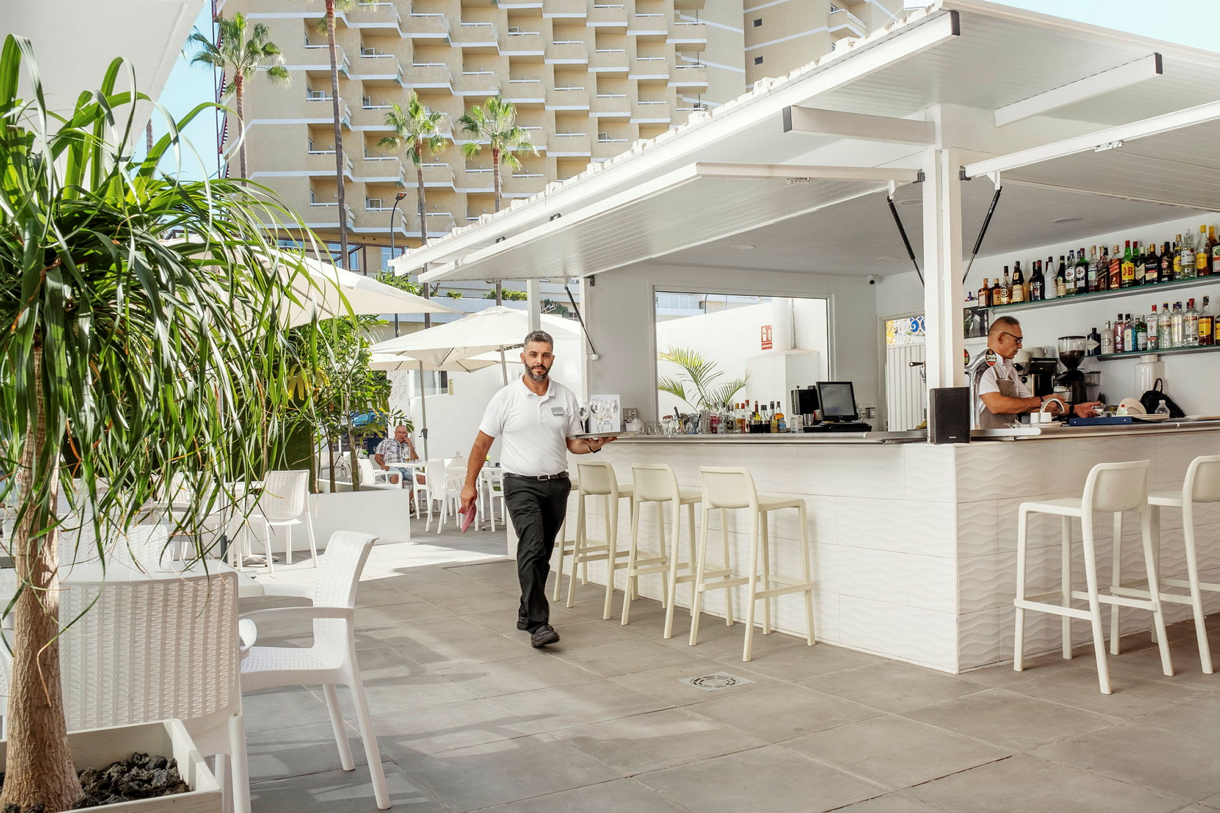 Kuva ulkobaarista, jossa istumapaikkoja, aurinkovarjoja ja rakennus taustalla. (Tekoälyn luoma teksti)