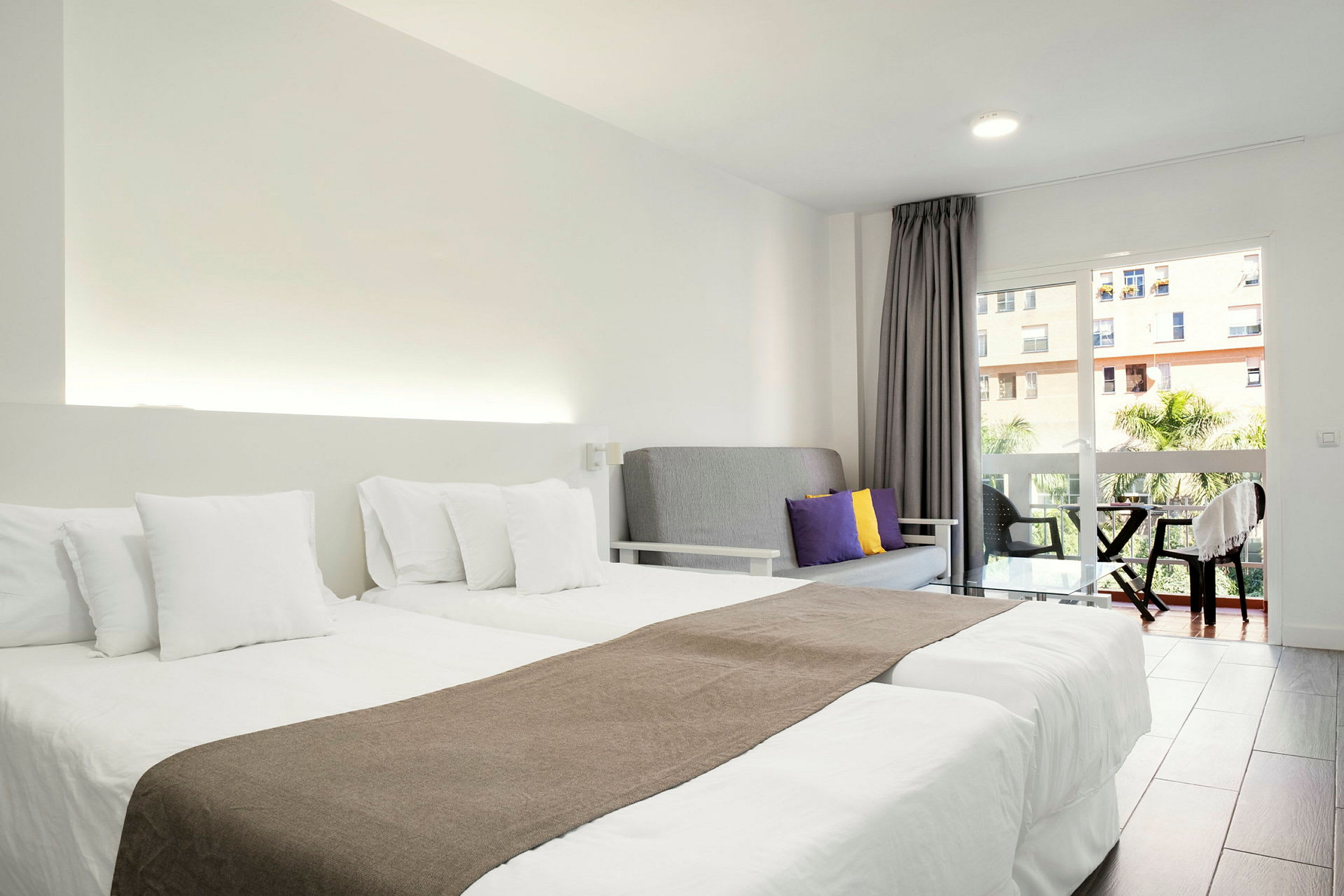 Hotellihuone, jossa on kaksi erillistä sänkyä valkoisin vuodevaattein, harmaa sohva värillisillä tyynyillä ja parveke näkymällä. (Tekoälyn luoma teksti)