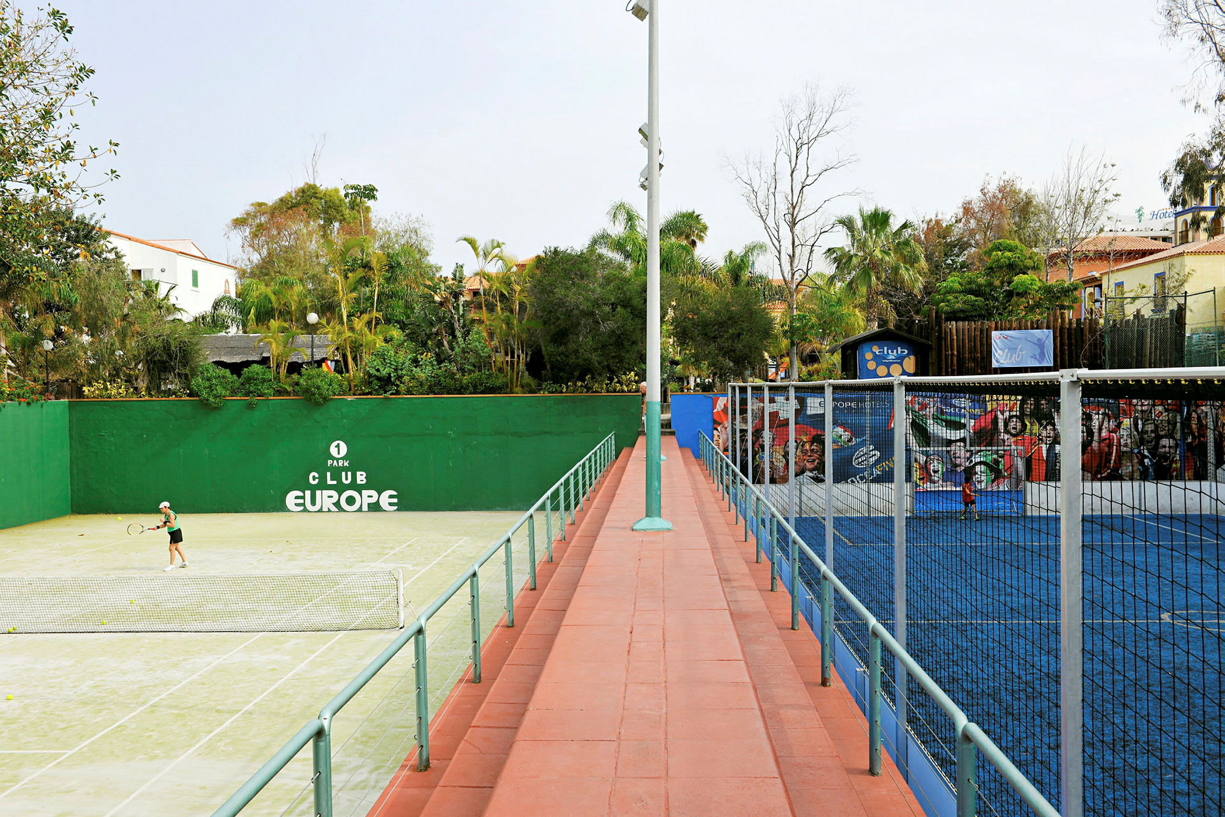 Vasemmalla vihreä tenniskenttä ja oikealla sininen padel-kenttä, ympäröitynä puilla ja rakennuksilla. (Tekoälyn luoma teksti)