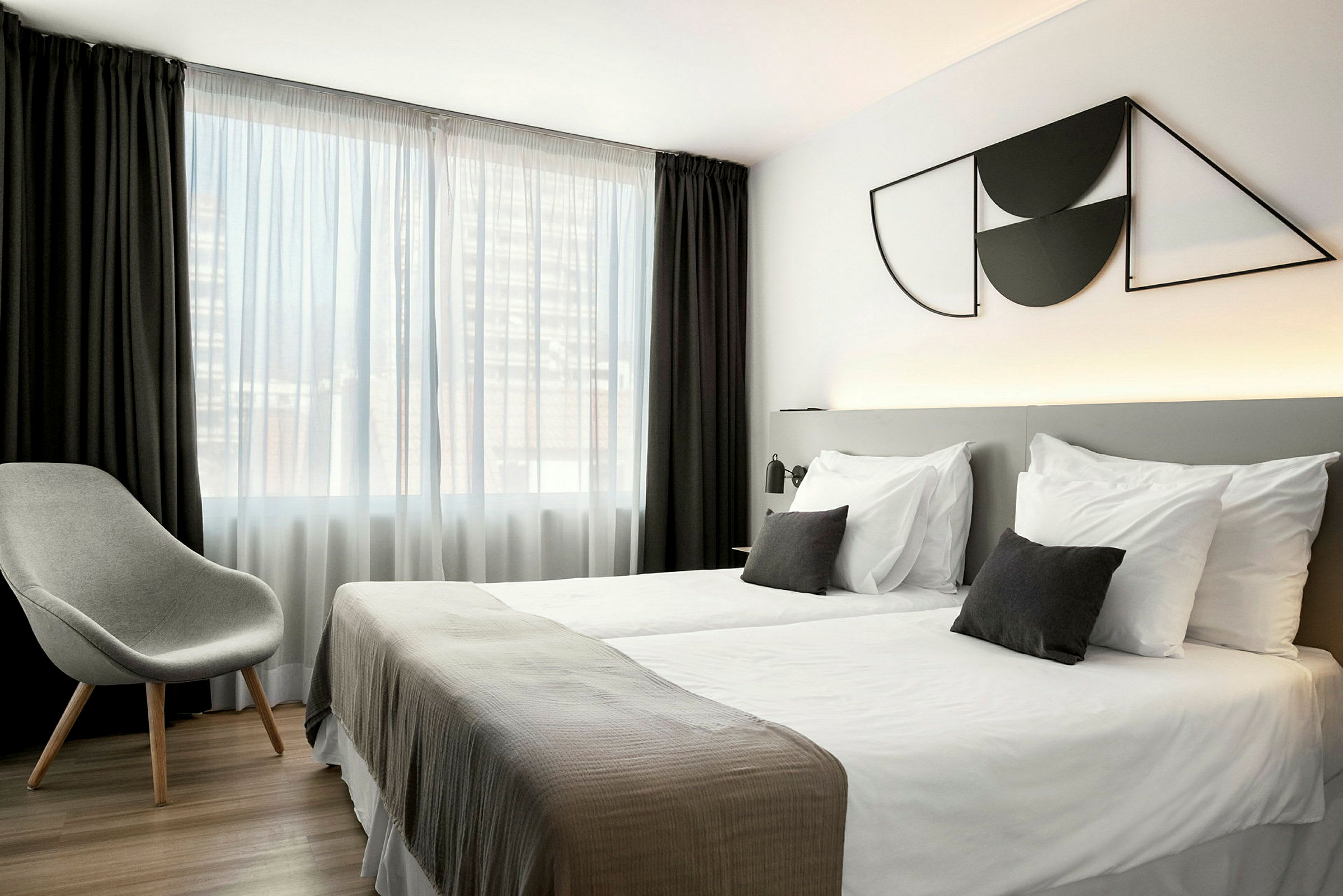 Hotellihuone, jossa on kaksi yhden hengen vuodetta, harmaa tuoli ja suuri ikkuna valkoisine ja tummine verhoineen. (Tekoälyn luoma teksti)