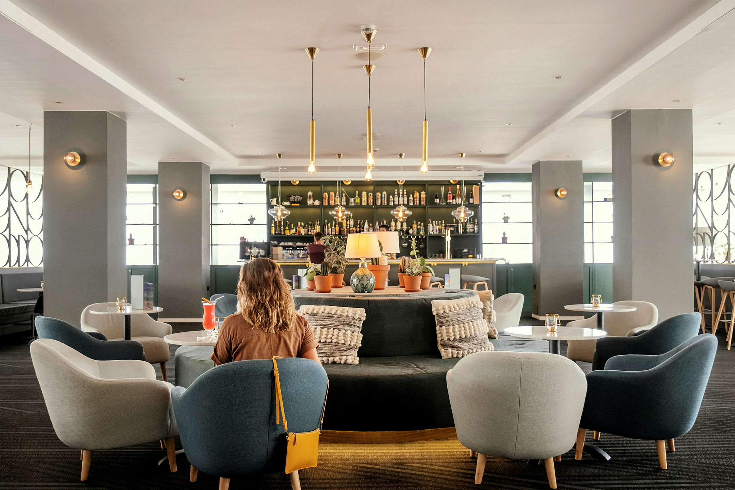 Modernin hotellibaarialueen sisätila istumapaikkojen ja koristeiden kanssa. Etualalla nainen selin kameraan. (Tekoälyn luoma teksti)