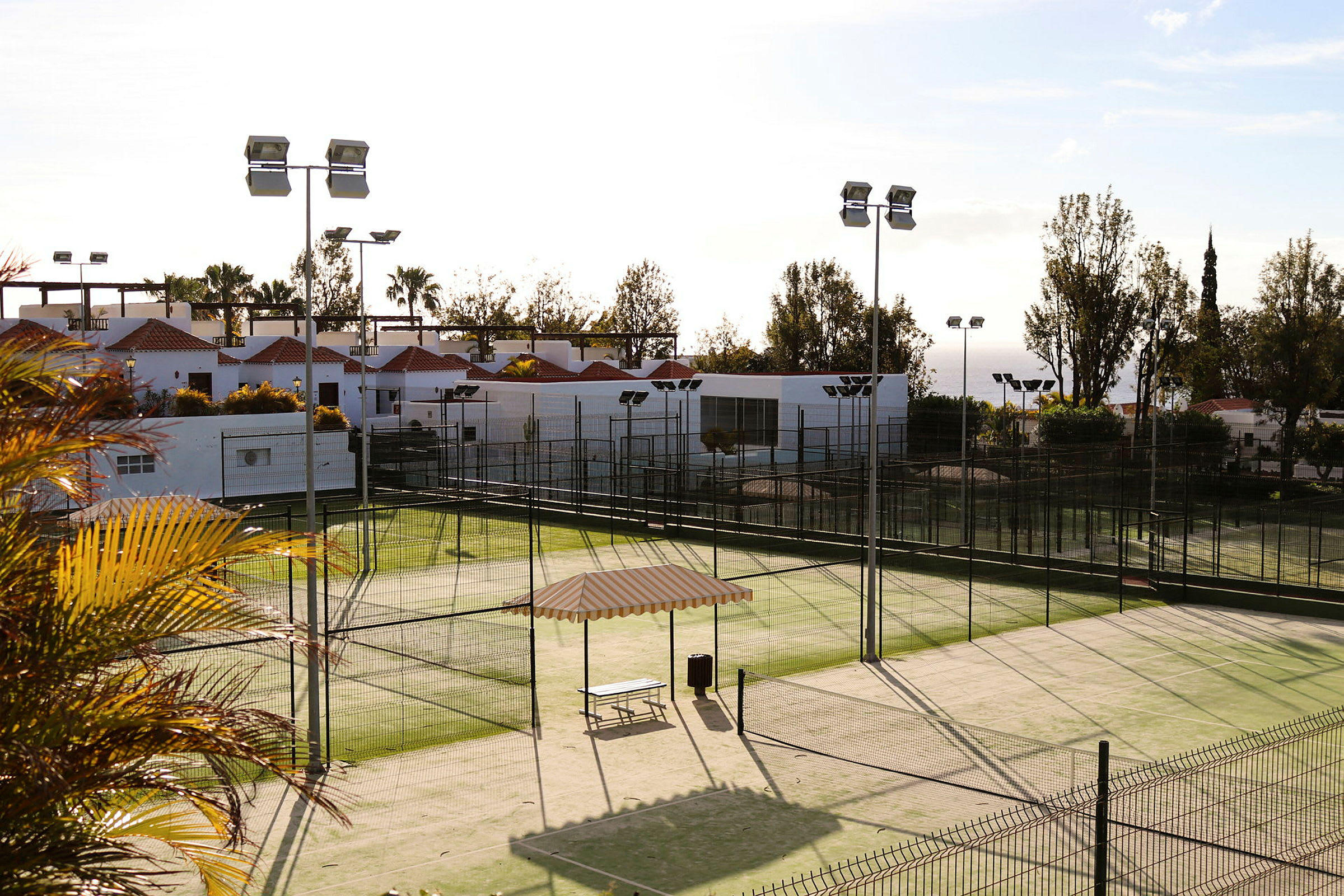 Kuva padeltenniskentästä, jota ympäröivät aidat. Taustalla näkyy valkoinen rakennus punaisilla katoilla. (Tekoälyn luoma teksti)