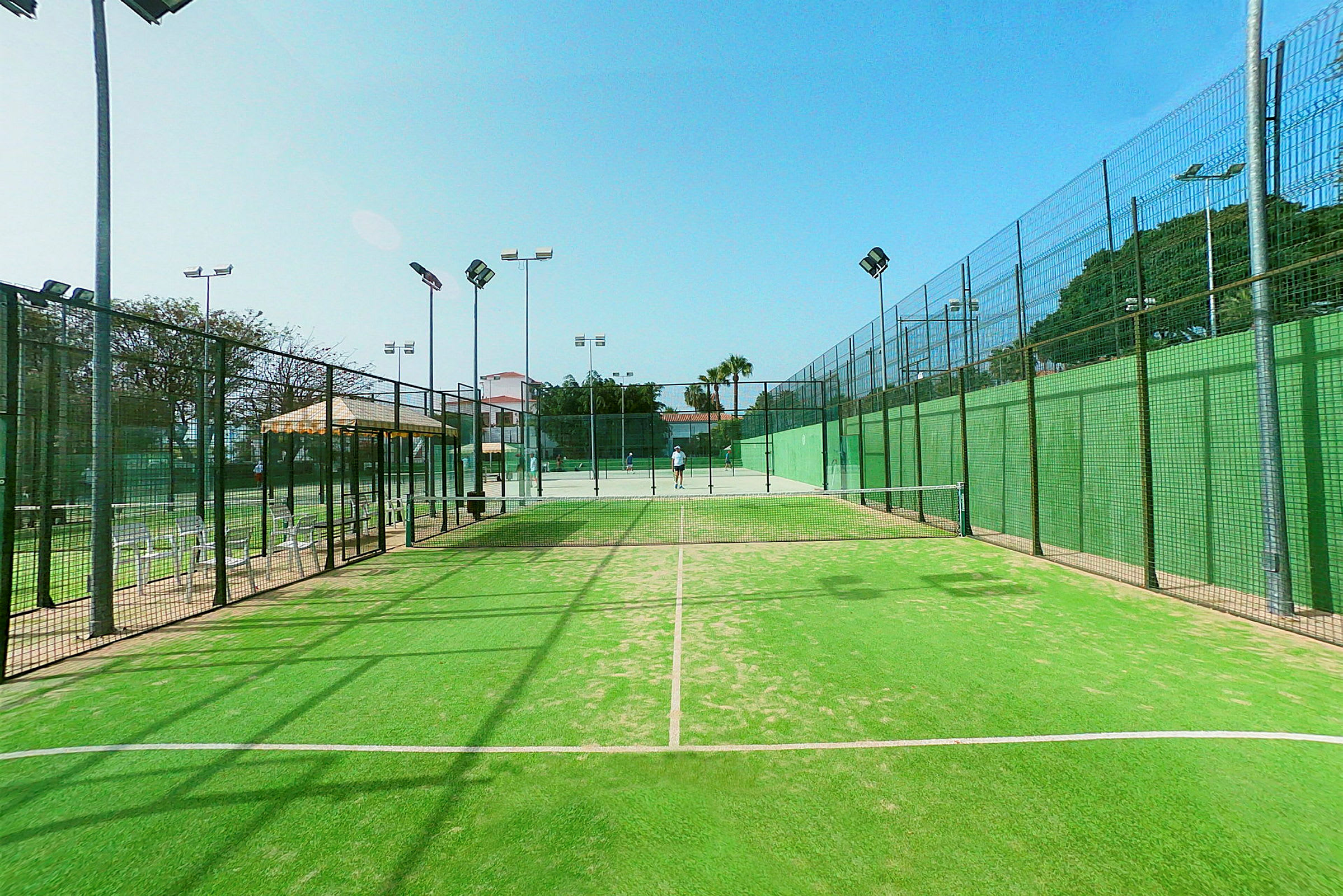 Vihreä padelkenttä, jossa verkot ja valaistus, ympäröimänä aidat. (Tekoälyn luoma teksti)
