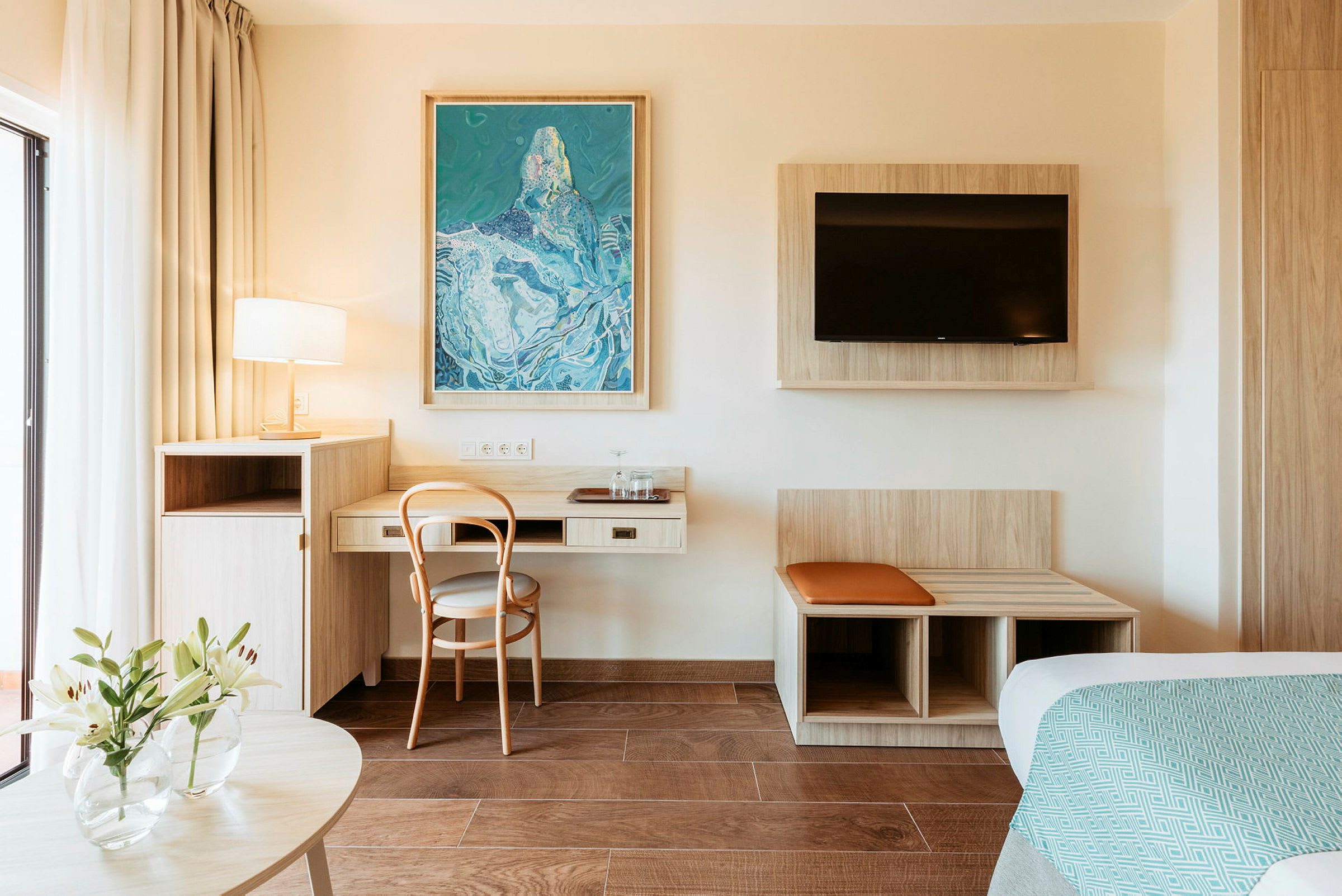 Hotellihuone, jossa on kirjoituspöytä, tuoli, seinään kiinnitetty televisio ja koristeellinen maalaus. (Tekoälyn luoma teksti)