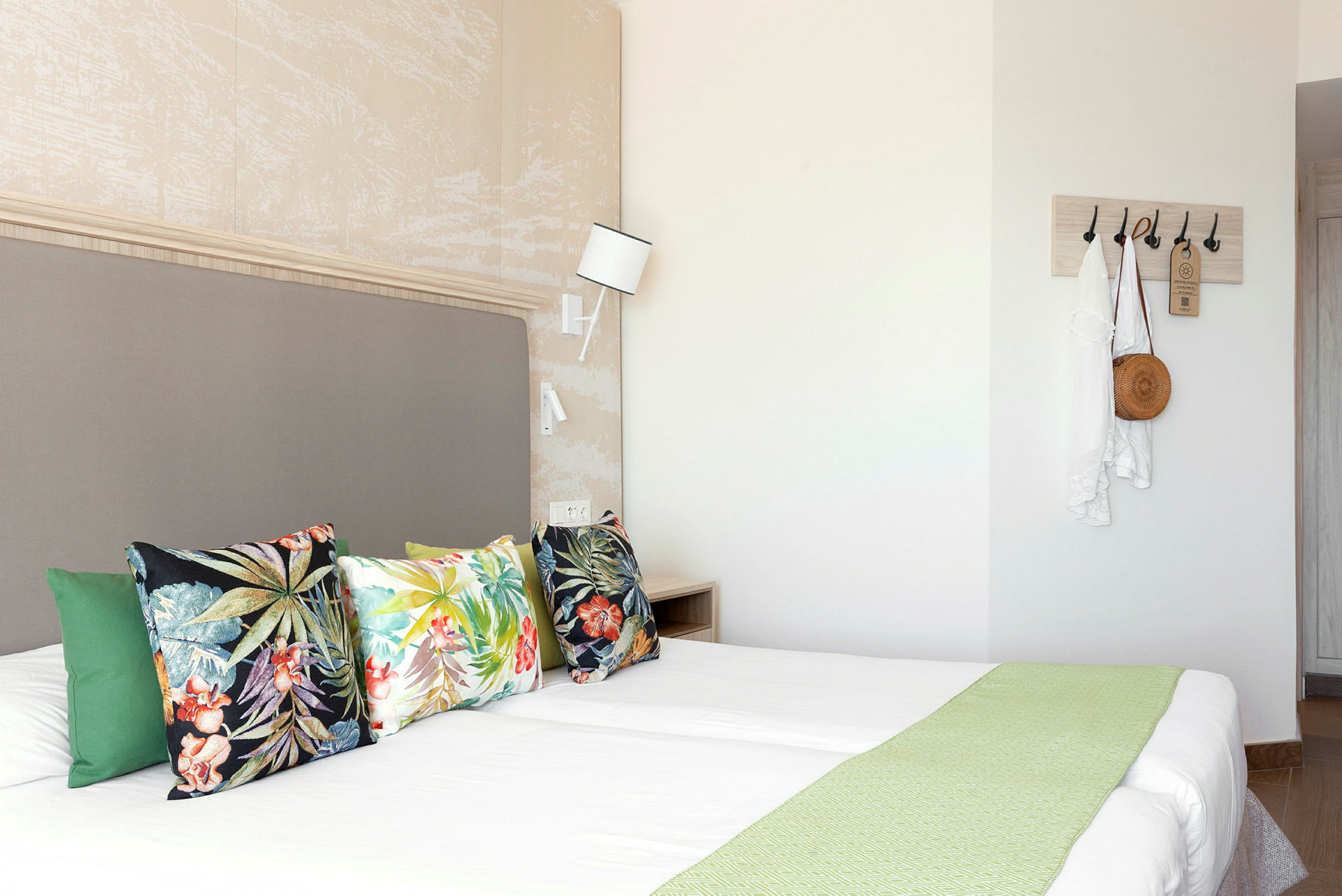 Hotellihuone, jossa parivuode, värikkäät tyynyt sekä seinäkoukkuja, joissa vaatteita ja laukku. (Tekoälyn luoma teksti)