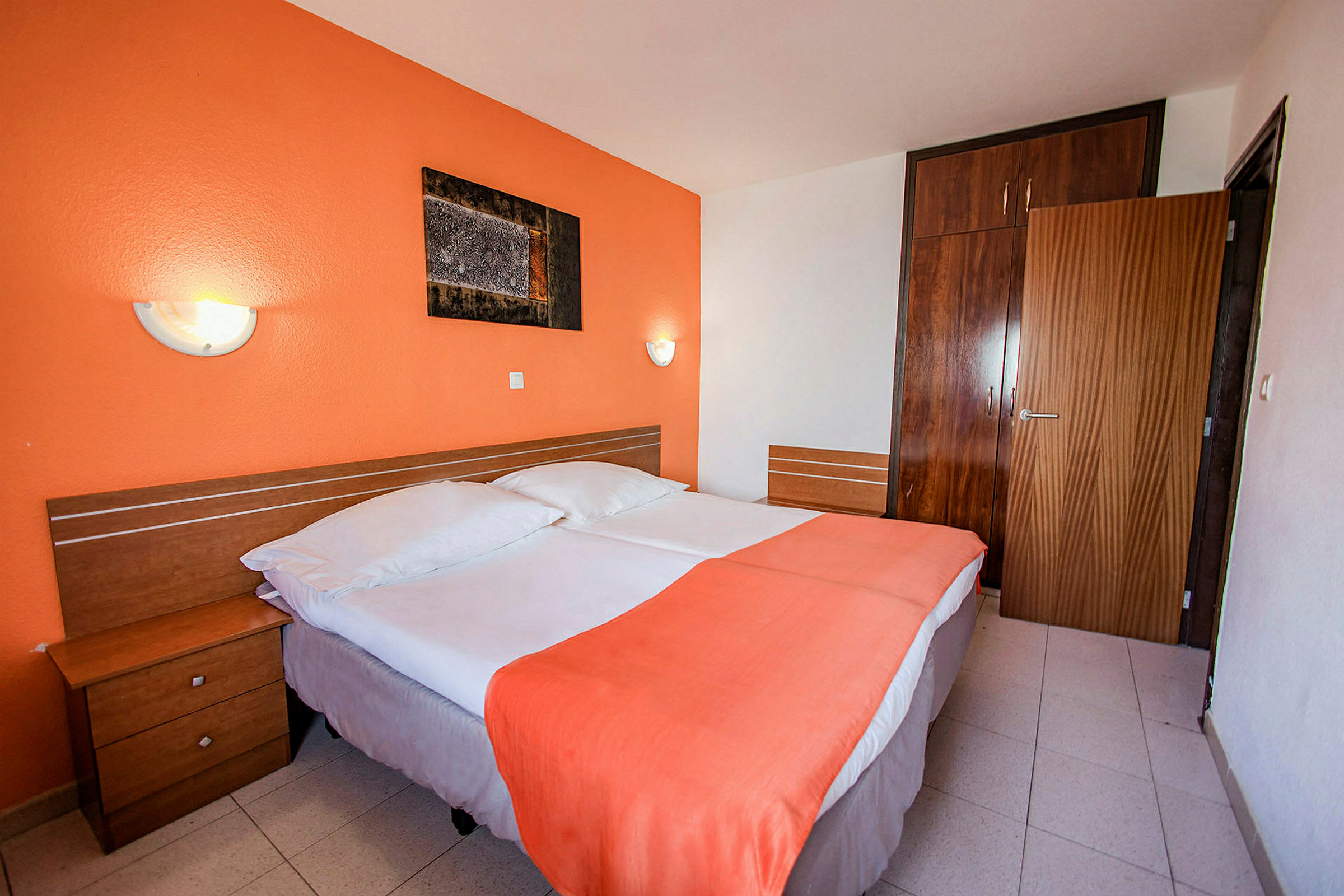 Hotellihuone, jossa kaksi yksittäistä sänkyä, oranssi seinä ja puukalusteet. (Tekoälyn luoma teksti)