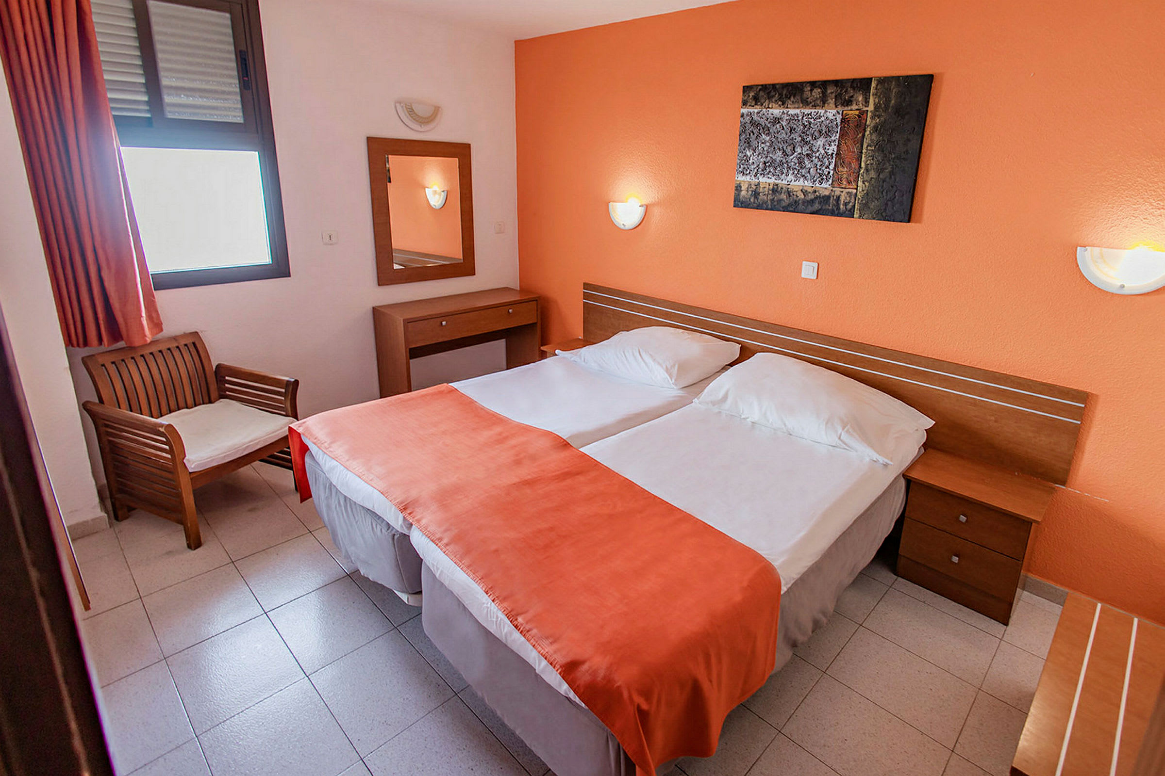 Hotellihuone, jossa on kaksi yhden hengen sänkyä oranssilla päiväpeitteellä, tuoli ikkunan vieressä ja peili seinälle kiinnitetyllä pöydällä. (Tekoälyn luoma teksti)