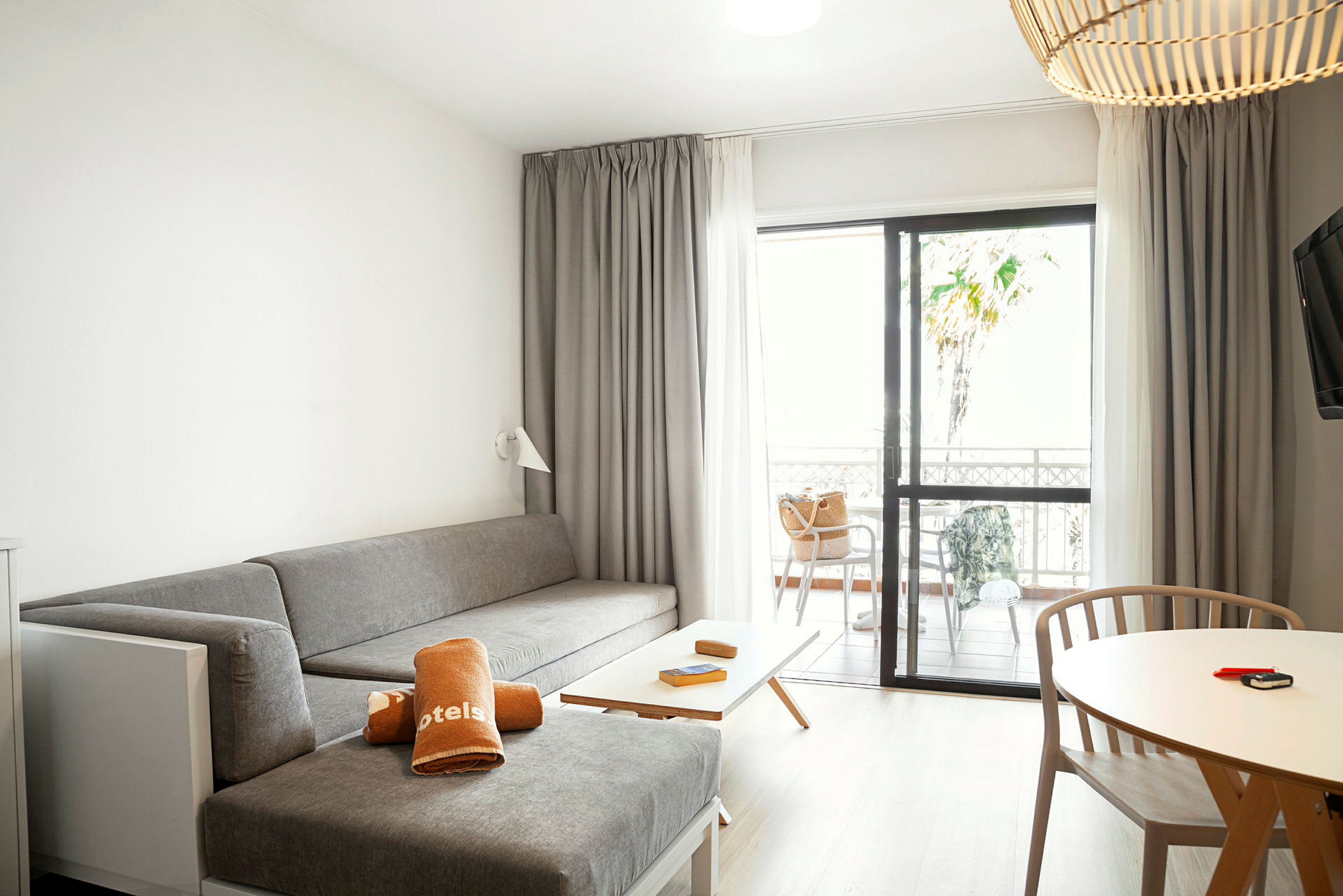 Hotellihuone, jossa on harmaa istuinalue, pöytä puutuoleineen ja terassi lasiovien läpi. (Tekoälyn luoma teksti)