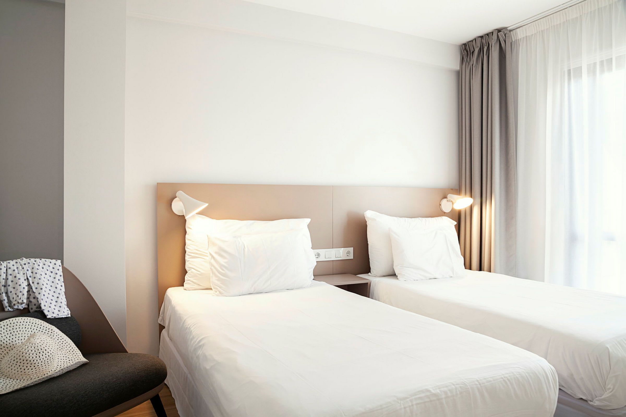 Hotellihuone, jossa on kaksi erillistä vuodetta, verhoilla varustettu ikkuna ja tuoli sivussa. (Tekoälyn luoma teksti)