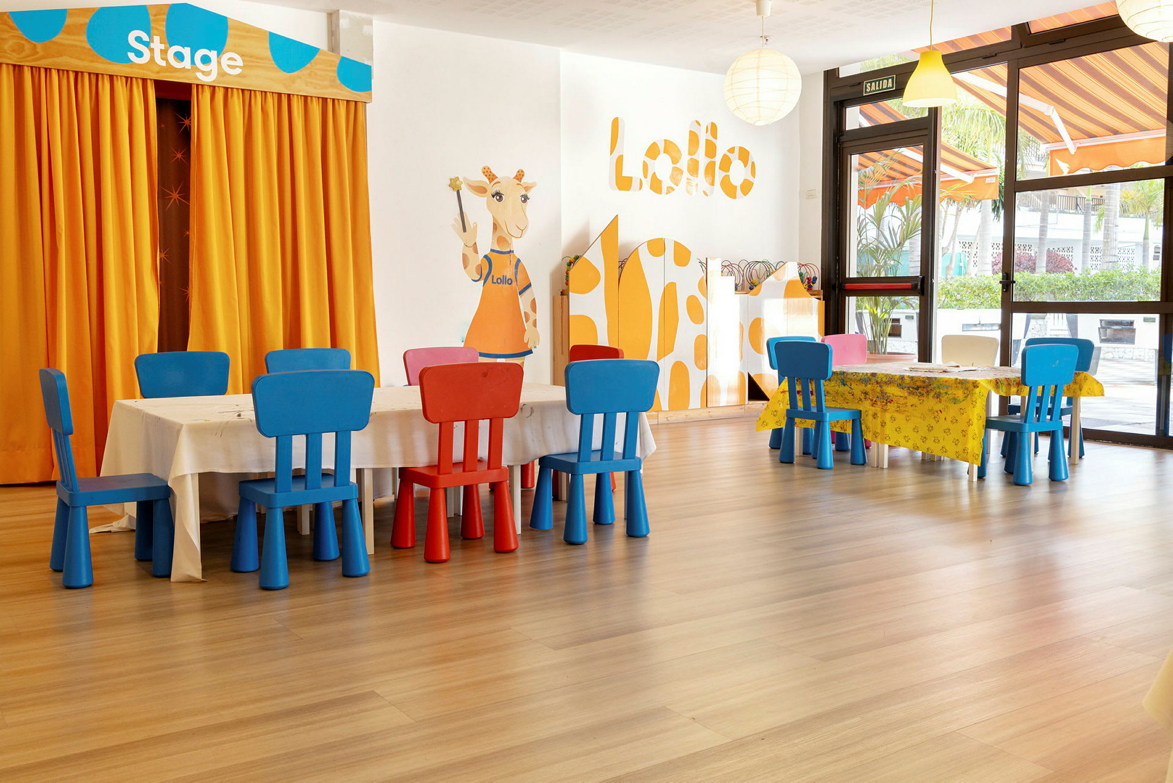 Lapsiystävällinen huone, jossa on pieniä pöytiä ja värikkäitä tuoleja. Kirahvin kuva oranssien verhojen vieressä, joissa lukee "Stage". (Tekoälyn luoma teksti)