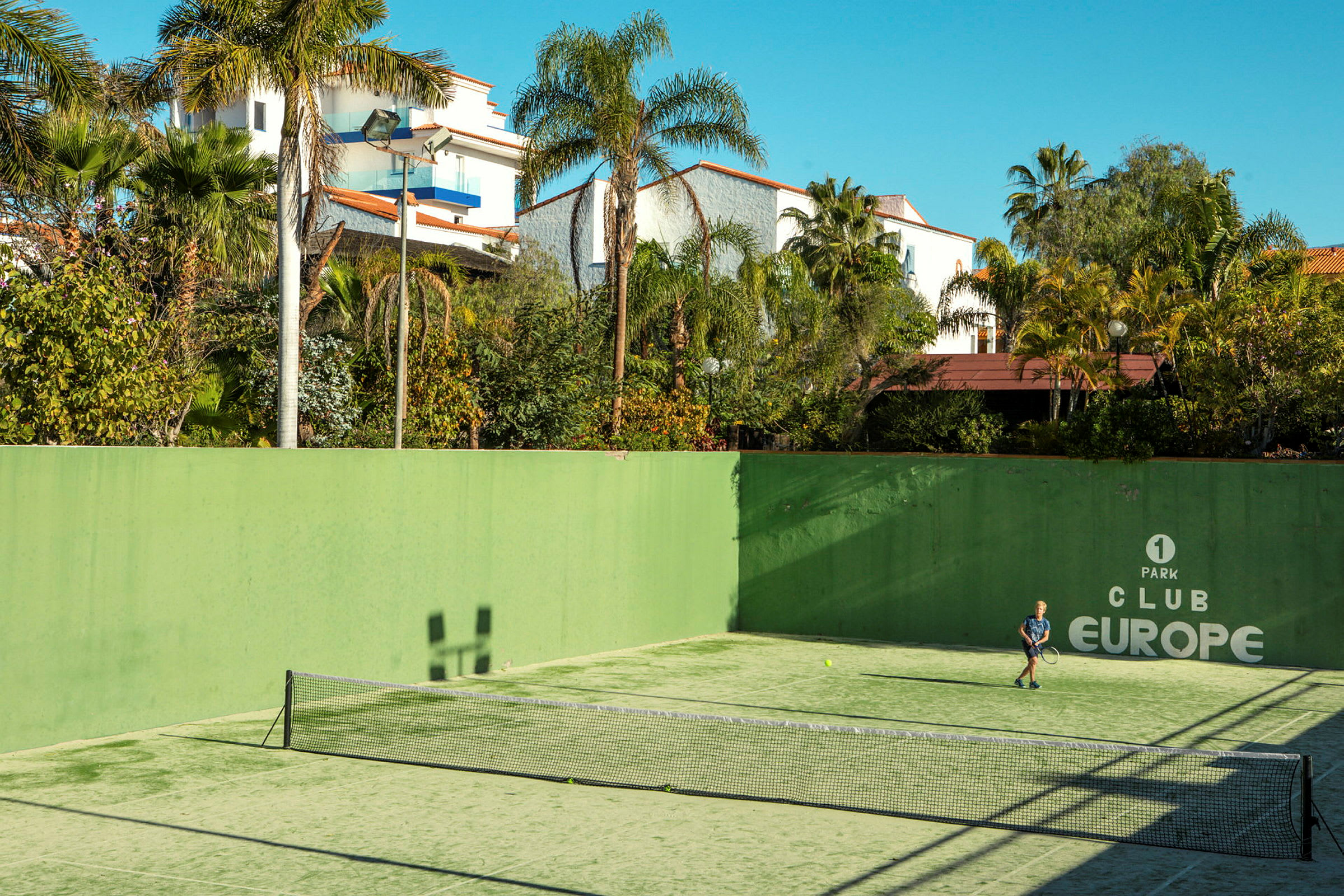 Ulkoilmatenniskenttä, jossa henkilö pelaa tennistä. Kenttää ympäröivät vihreät kasvit ja rakennukset. (Tekoälyn luoma teksti)