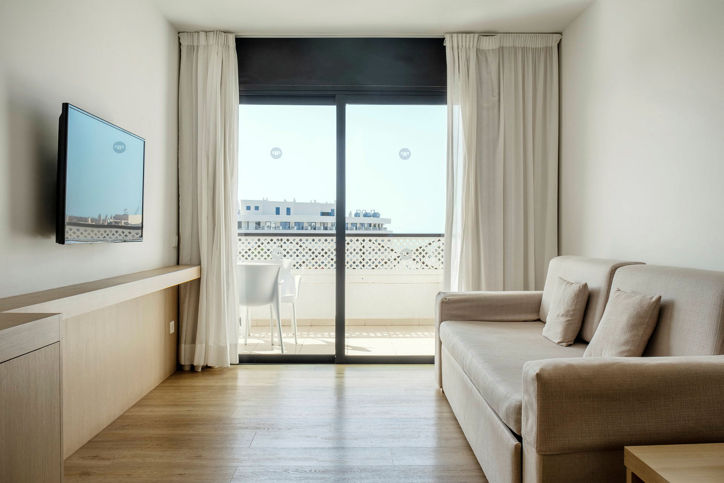 Valoisa ja moderni hotellihuone, jossa on sohva, seinälle kiinnitetty TV ja suuri ikkuna, josta avautuu näkymä parvekkeelle. (Tekoälyn luoma teksti)