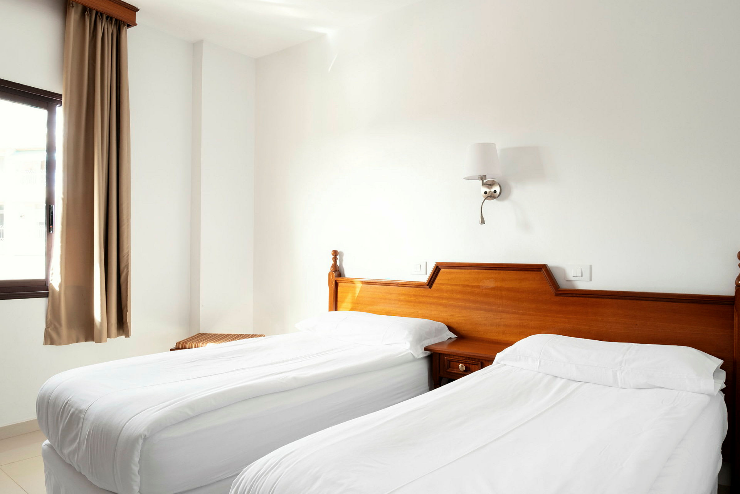 Hotellihuone, jossa on kaksi yksittäissänkyä valkoisine vuodevaatteineen, puukalusteita ja ikkuna, jossa on beigessäverhot. (Tekoälyn luoma teksti)