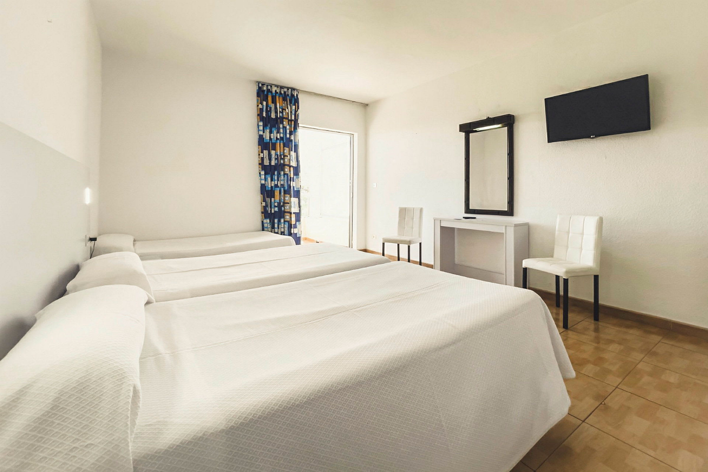 Hotellihuone, jossa on kolme yhden hengen sänkyä, työpöytä peilillä, kaksi tuolia ja seinään kiinnitetty televisio. (Tekoälyn luoma teksti)