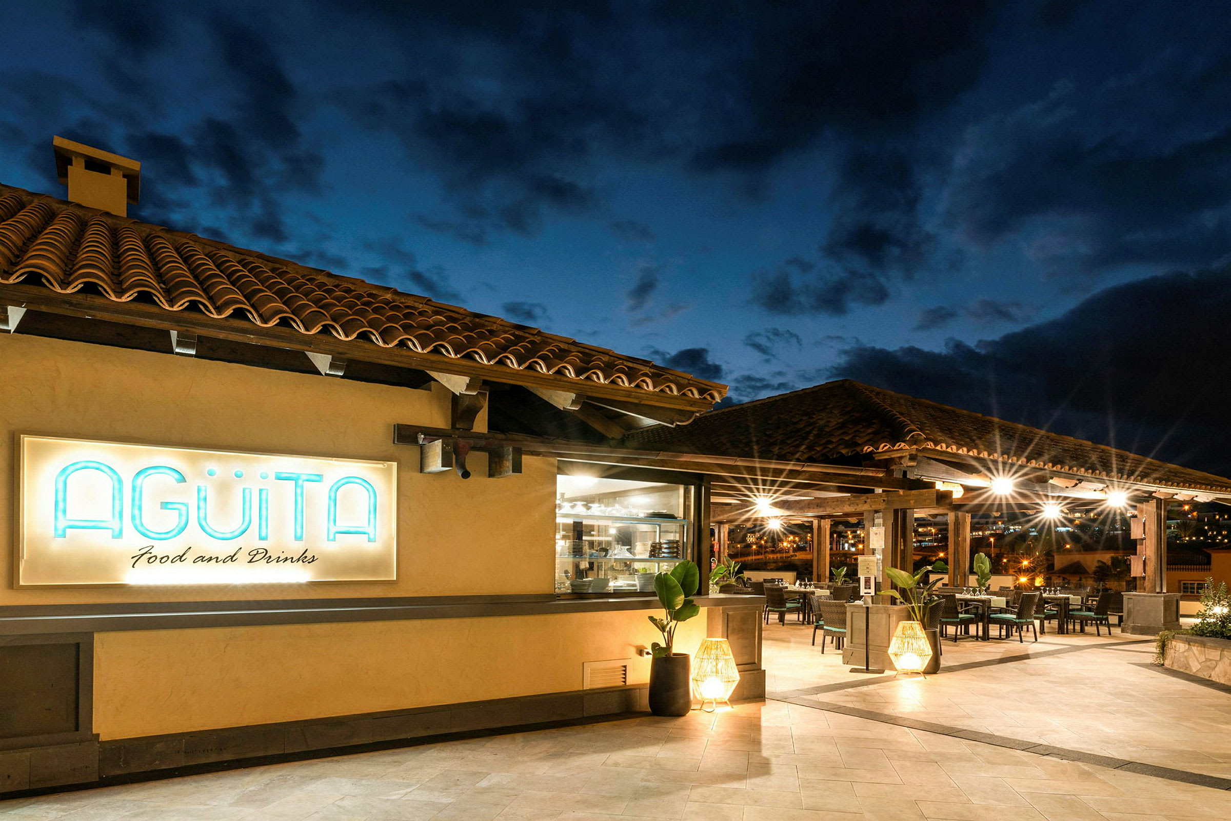 Ravintolan ulkokuva, jossa tiilikatto ja valaistu Agüita-kyltti. (Tekoälyn luoma teksti)