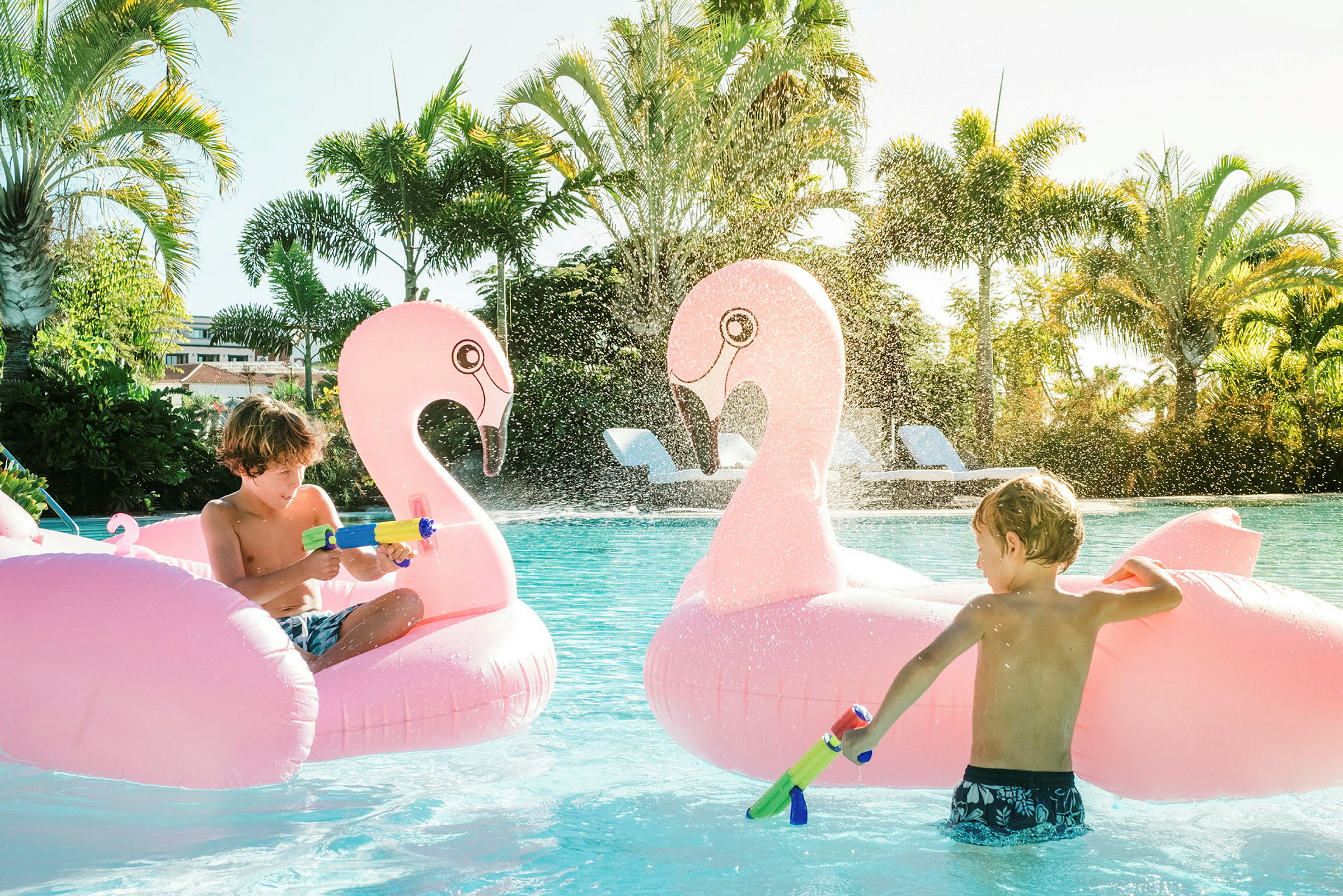 Kaksi puhallettavaa flamingohahmoa altaassa, vettä loiskuu ja ympärillä palmuja. (Tekoälyn luoma teksti)