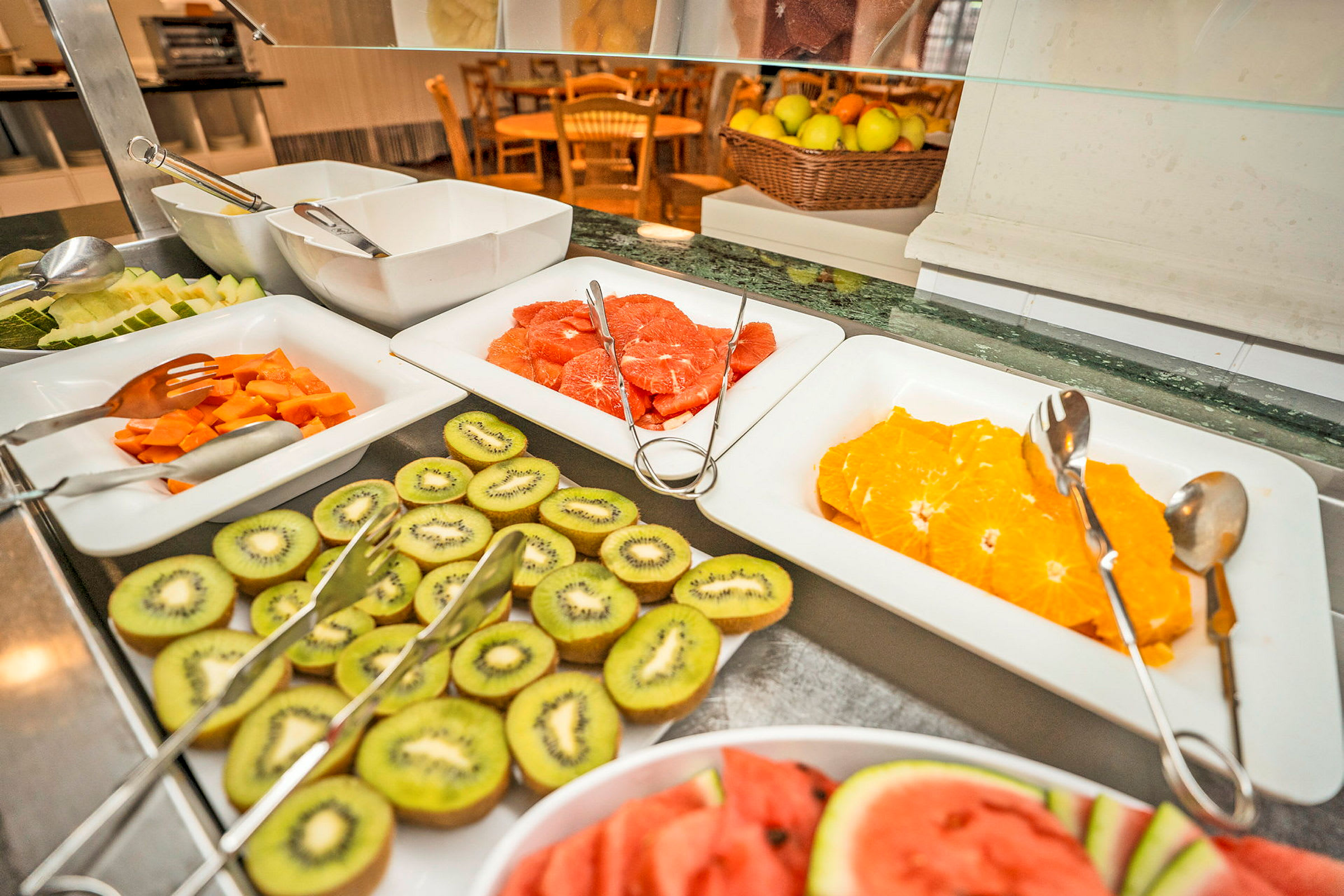 Buffetpöytä, jossa on viipaloituja hedelmiä, kuten appelsiineja, kiivejä ja vesimeloneja vadeissa. (Tekoälyn luoma teksti)